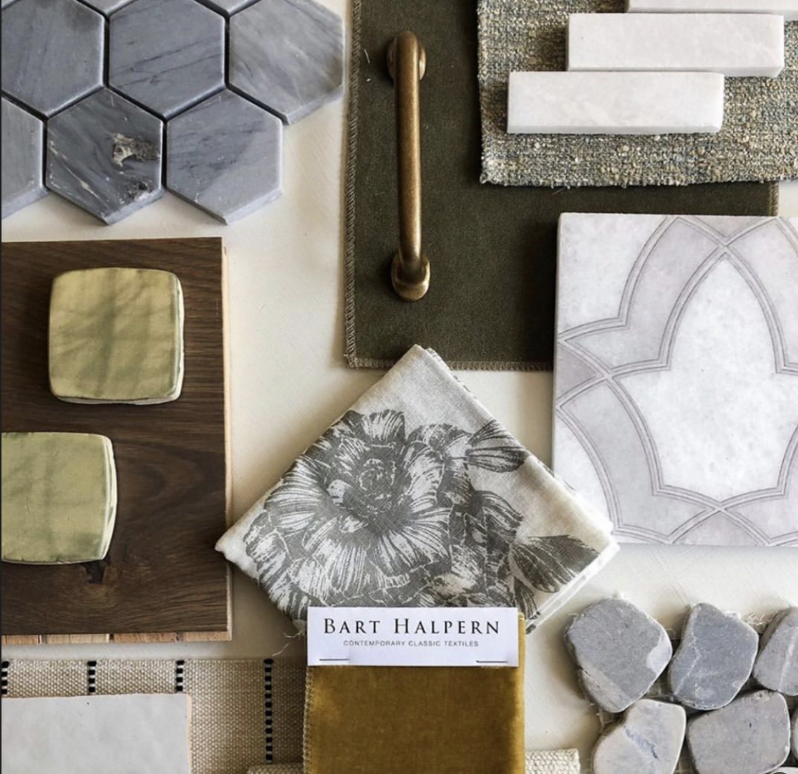 A selection of interior design materials including hexagonal marble tiles, a candlestick holder on a fabric swatch, a fabric with a floral pattern, a white tile with an arch pattern, three gray heart-shaped stones, a piece of velvet fabric, and a small wooden table with green square upholstered cushions.