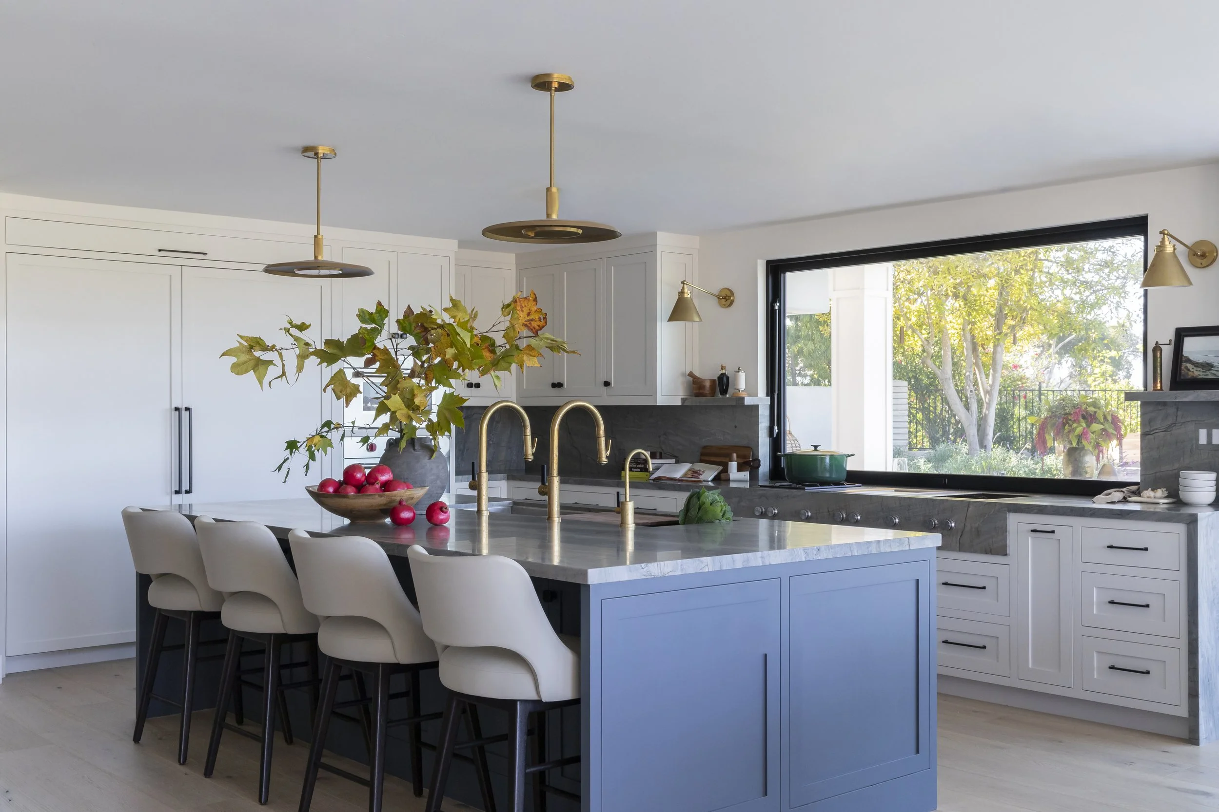 Modern kitchen with white cabinets, a blue island, gold fixtures, and a large window overlooking a garden.