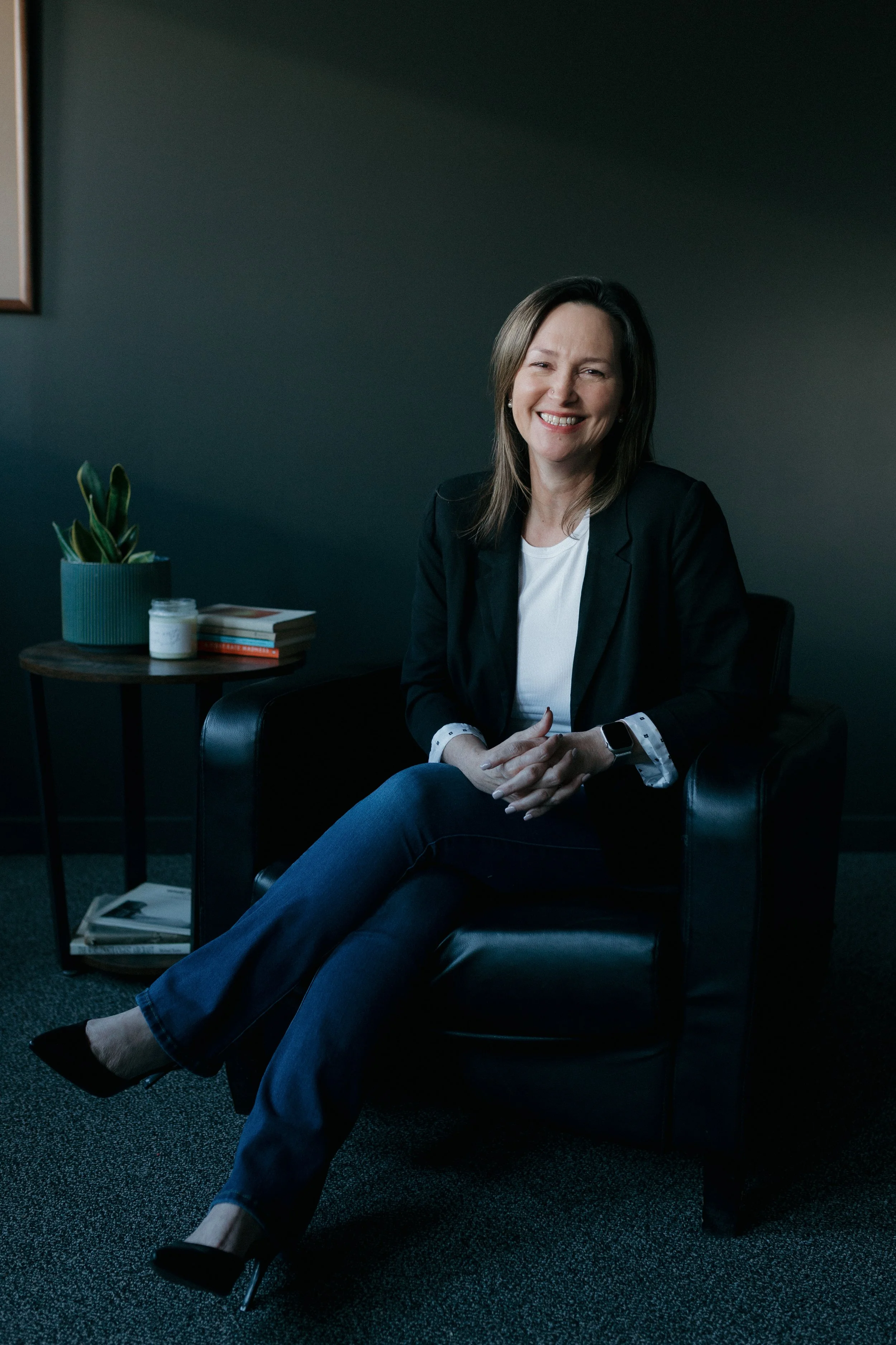 A woman with shoulder-length brown hair, wearing a black blazer, white shirt, and dark jeans, sitting in a black armchair with her legs crossed and smiling in a dark room. There are some books, a plant, and a white jar on a side table next to her.