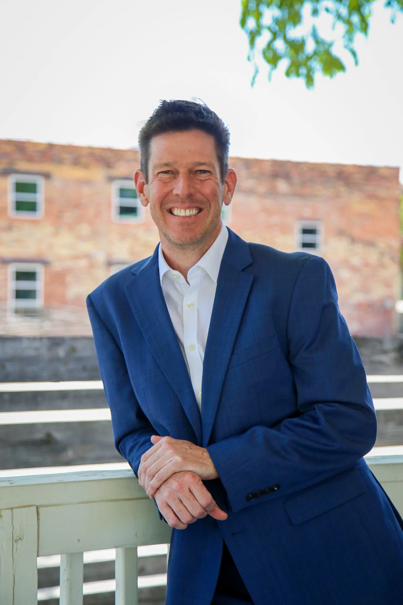 A man in a blue suit and white shirt leaning on a porch railing outdoors, smiling at the camera with a brick building in the background.