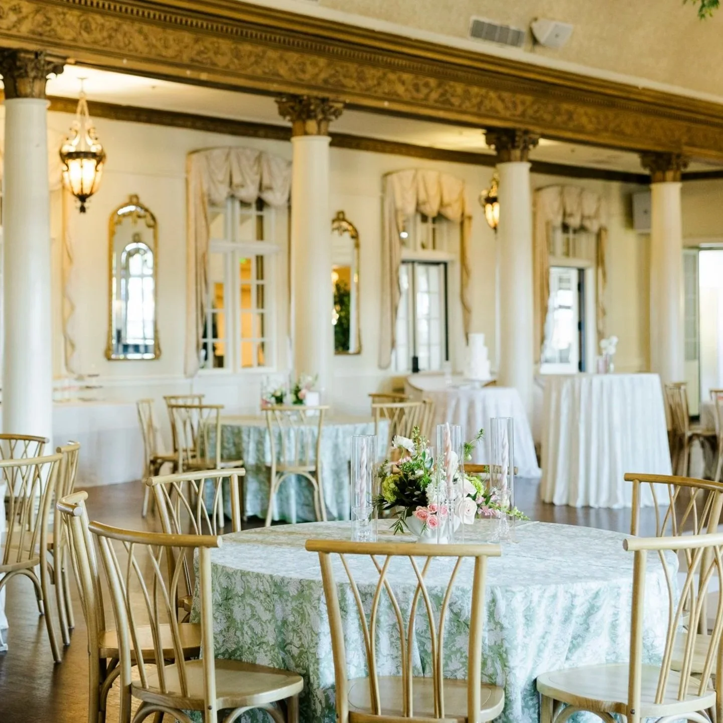 Elegant banquet room decorated for a celebration with round tables draped in floral tablecloths, floral centerpieces, chairs, chandeliers, large windows, and curtains.
