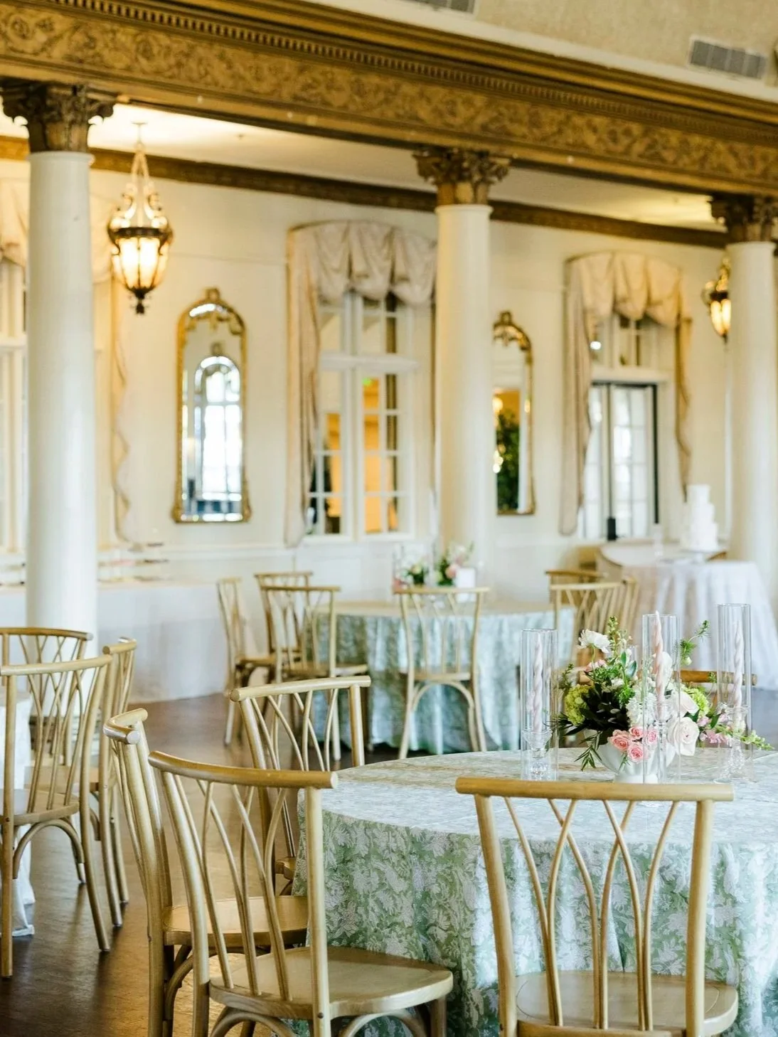 Elegant banquet hall with round tables covered in floral tablecloths, surrounded by wooden chairs, and decorated with flower centerpieces. The room features large windows with draped curtains, ornate wall mirrors, and classic pendant lights.
