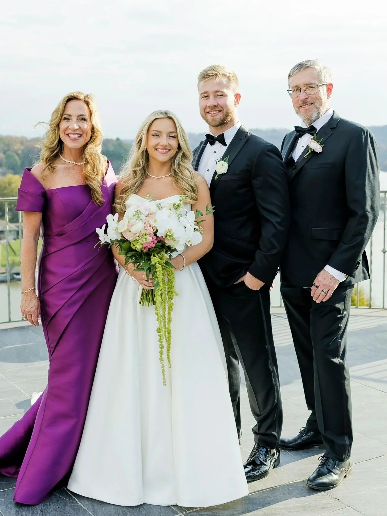 A wedding portrait of a woman in a white dress holding a bouquet, with three people in formal attire (two men and one woman) standing outdoors on a balcony, smiling.
