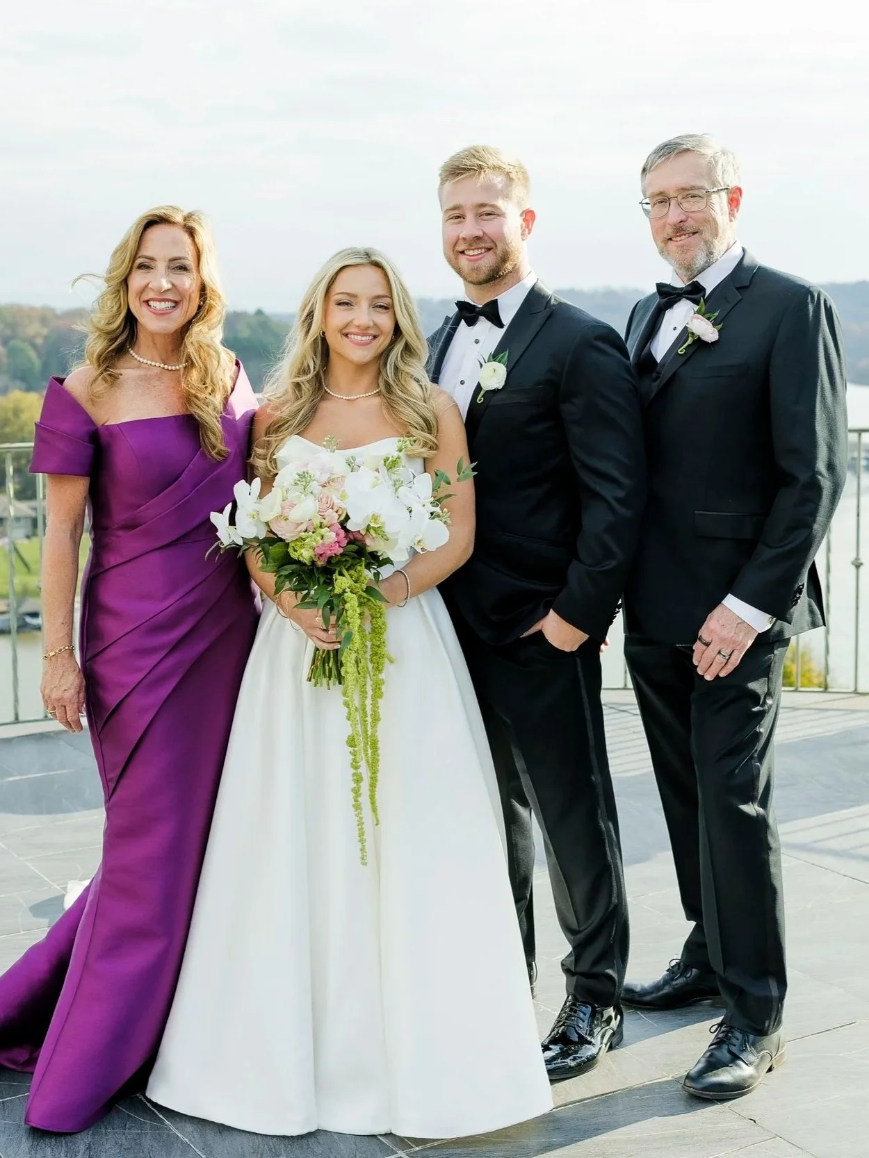 A group of four people dressed in formal wedding attire smiling outdoors, with a scenic background of water and trees. A bride holds a bouquet of flowers, flanked by two men in tuxedos and a woman in a purple gown.