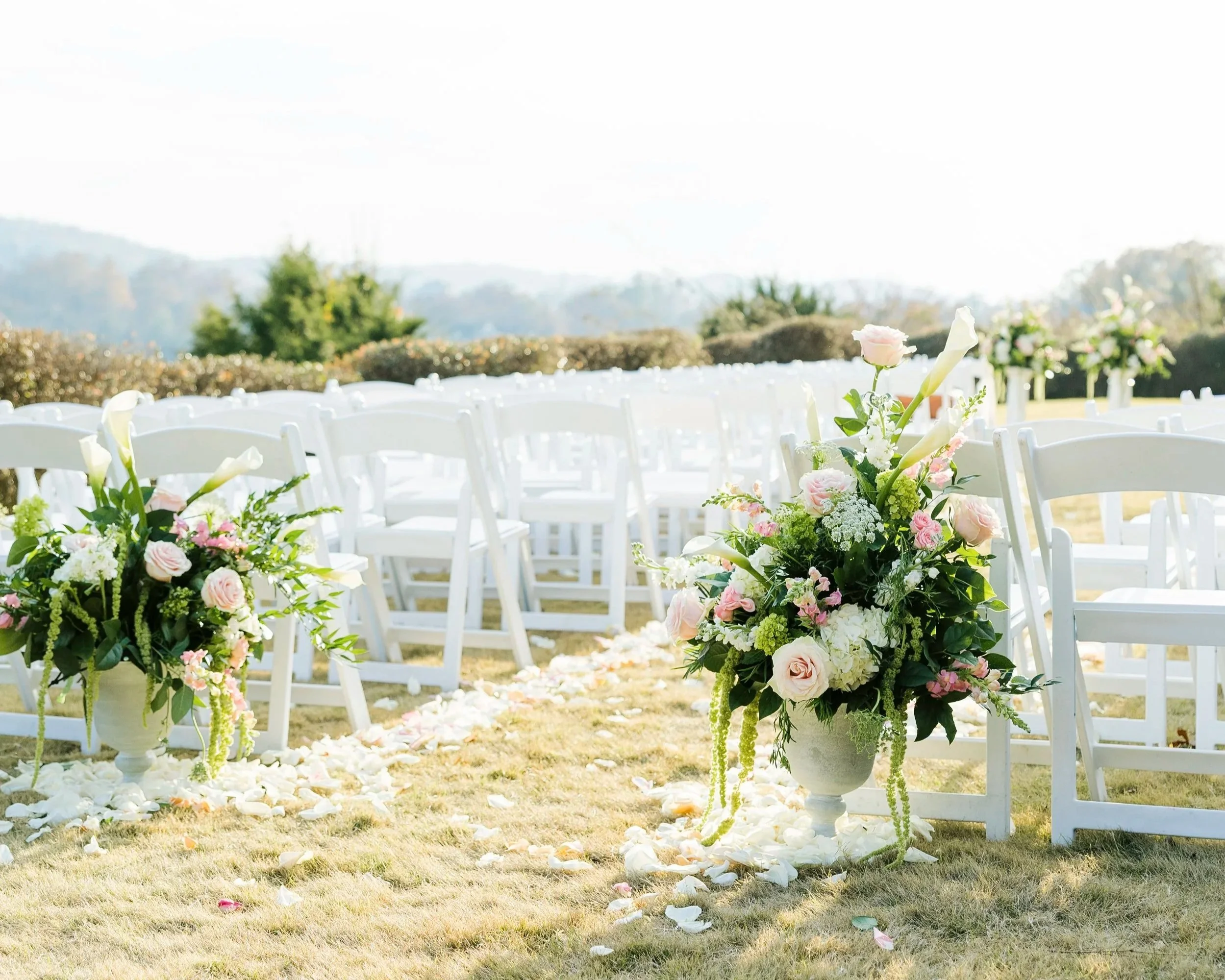 Outdoor wedding ceremony setup with white chairs, floral arrangements in white vases with pink and white flowers, and rose petal decorations on the grass aisle.