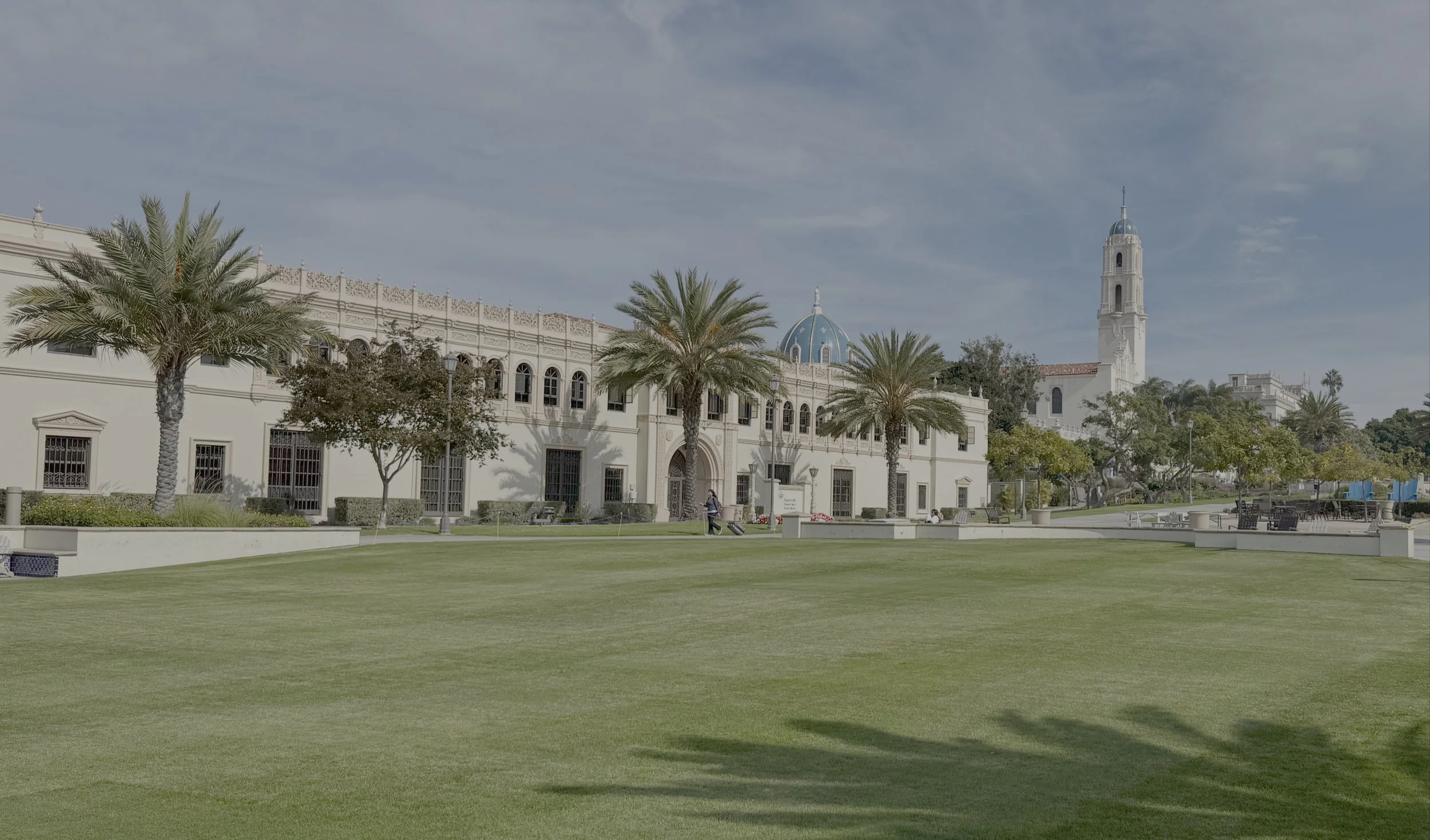 A large white building with arched windows and a blue domed roof, surrounded by palm trees and a well-maintained green lawn with patio furniture and a few people walking nearby, under a partly cloudy sky.