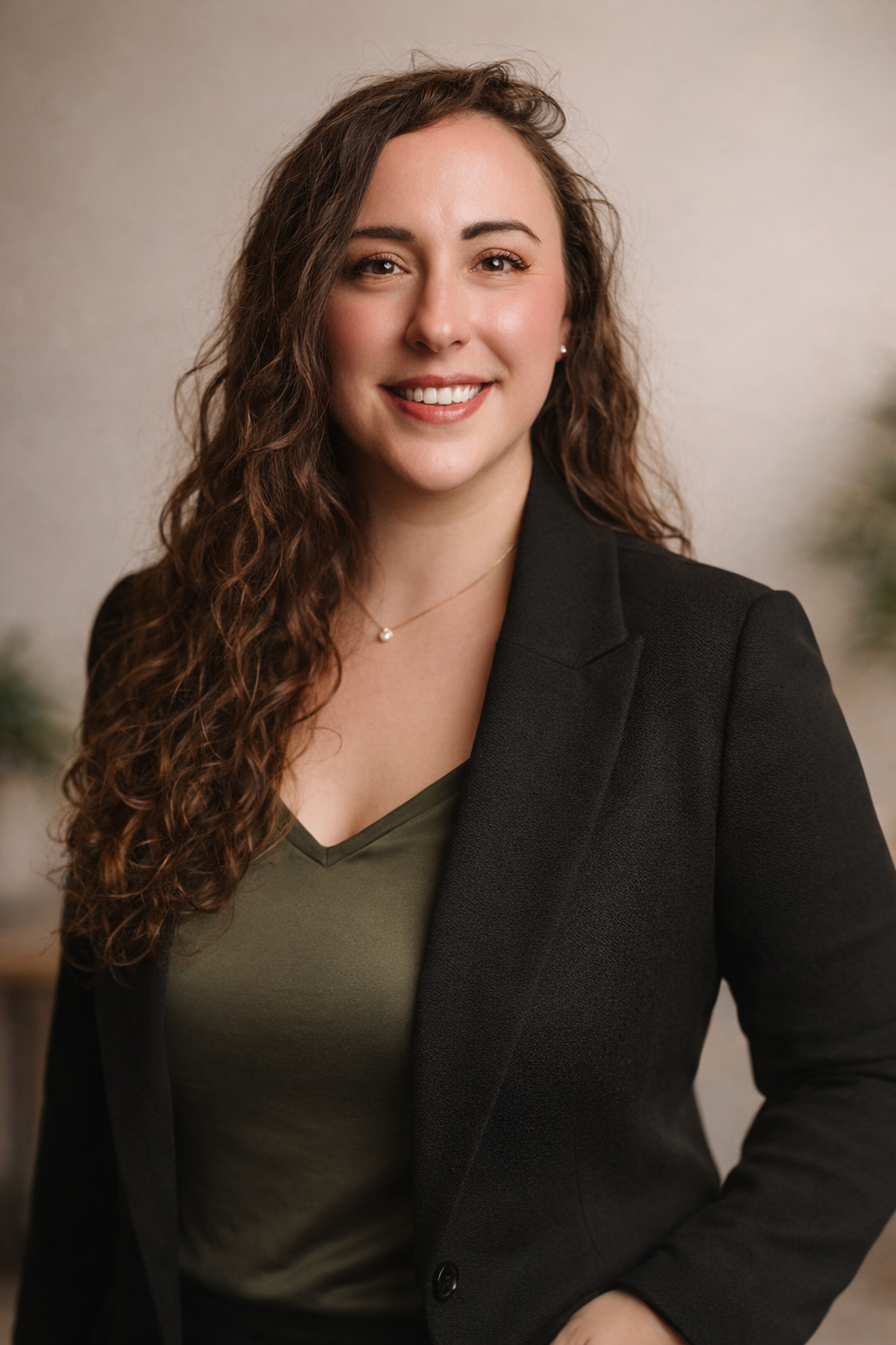 An attractive woman with long curly brown hair, wearing a black blazer over an olive green top, smiling at the camera against a neutral background.