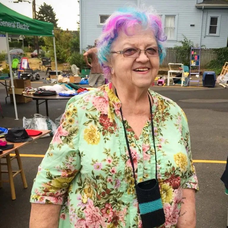 An elderly woman with colorful pastel hair, glasses, and a floral shirt, smiling at an outdoor event with tables and various items in the background.
