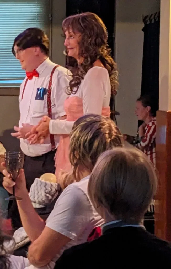 A woman with long, curly brown hair wearing a peach and white dress stands next to a man with glasses, dark hair, and a red bow tie, dressed in a white shirt with suspenders and pens in the pocket, in a room with multiple people, some of whom are seated at a table.
