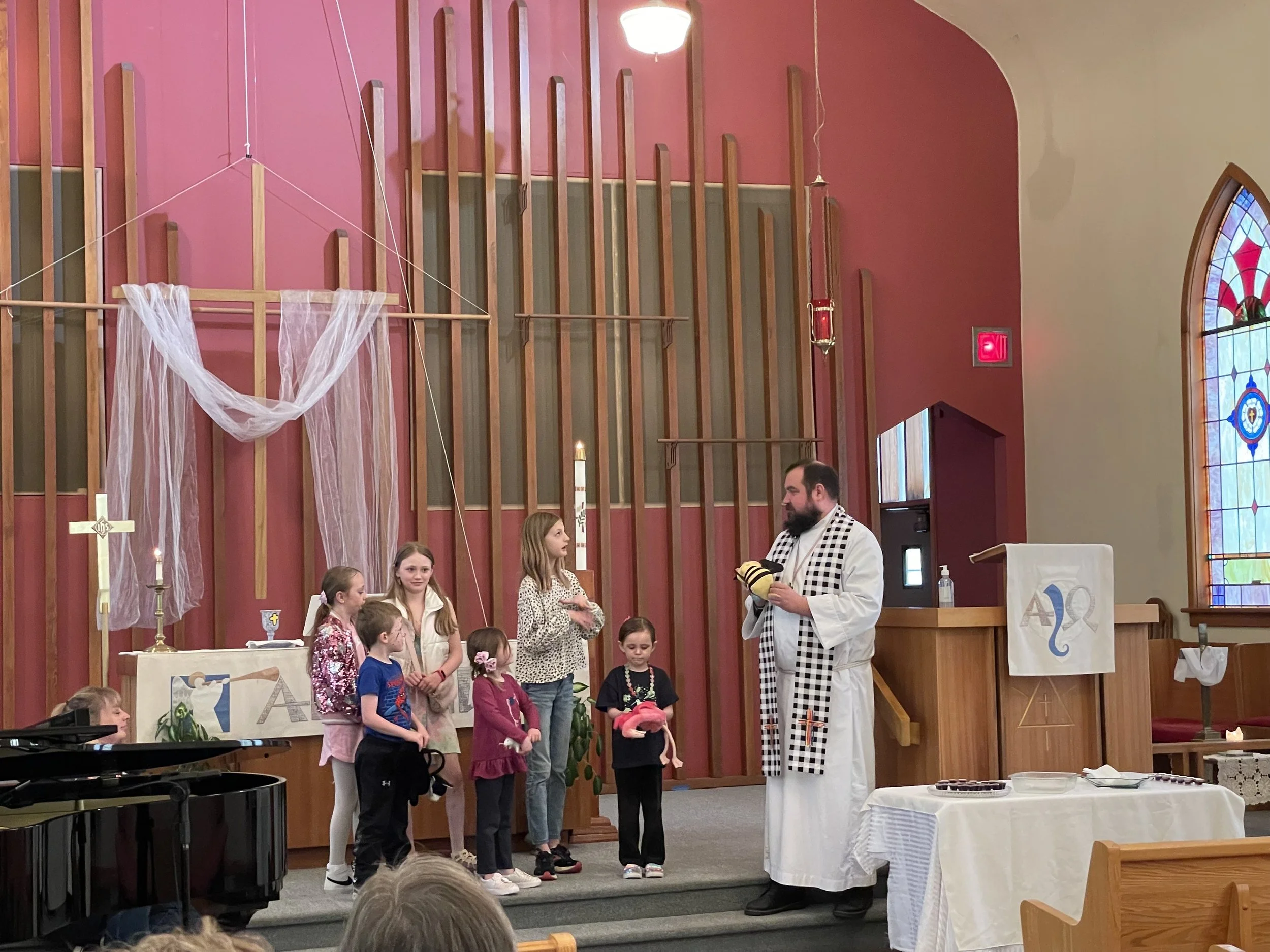 Children standing in a line at a church altar during a religious service, with a priest holding a stole, inside a church with stained glass windows and a wooden backdrop.