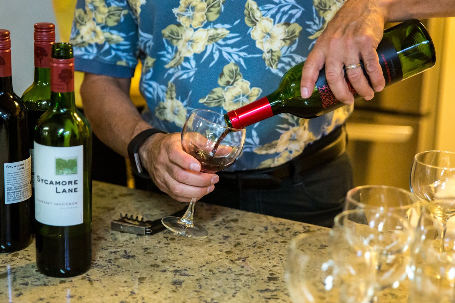 Person pouring red wine into a wine glass from a green bottle labeled Sycamore Lane, with several other wine bottles and empty glasses on a kitchen countertop.