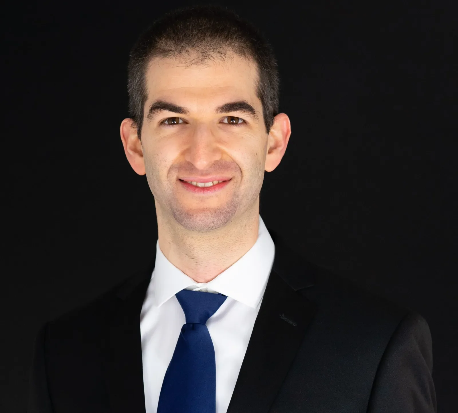 Professional headshot of a man in a black suit, white shirt, and blue tie, smiling against a black background.