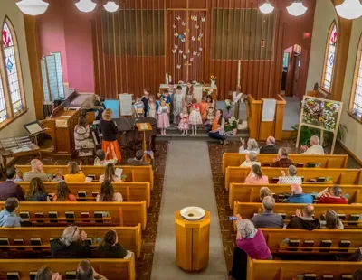 Children performing at a church service with a piano and choir, audience seated on wooden pews, decorated with butterflies and stained glass windows.