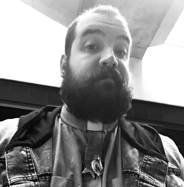 A man with a beard and slicked-back hair taking a selfie. He is wearing a clerical collar, a badge, and a denim jacket, and is in an indoor setting.