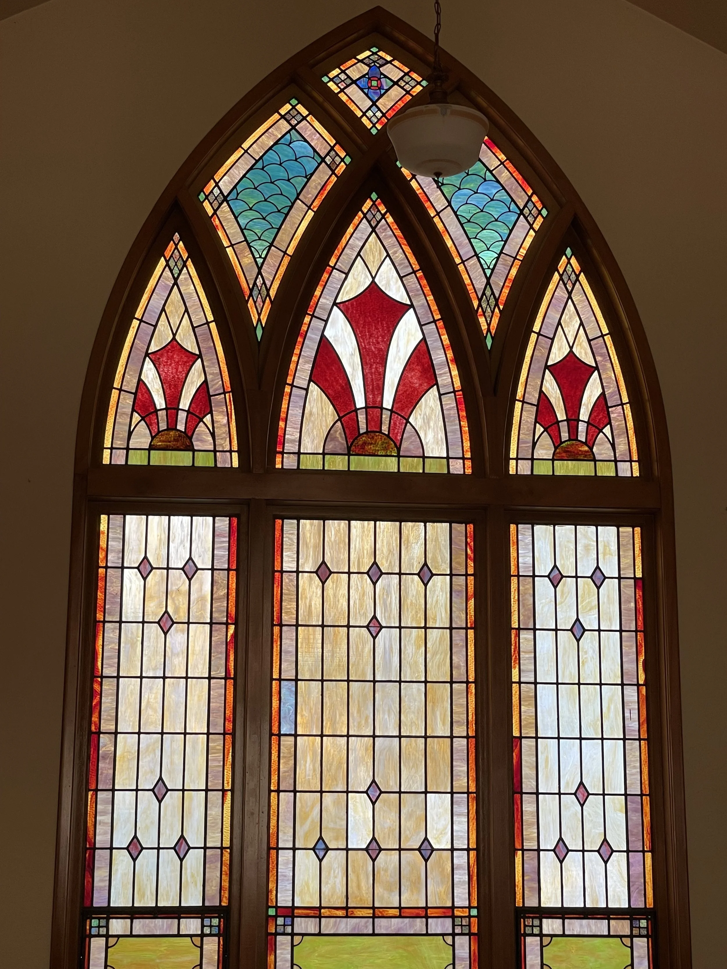 Colorful stained glass window with geometric patterns and arches, featuring red, blue, yellow, and purple hues, and a hanging light fixture in the center.