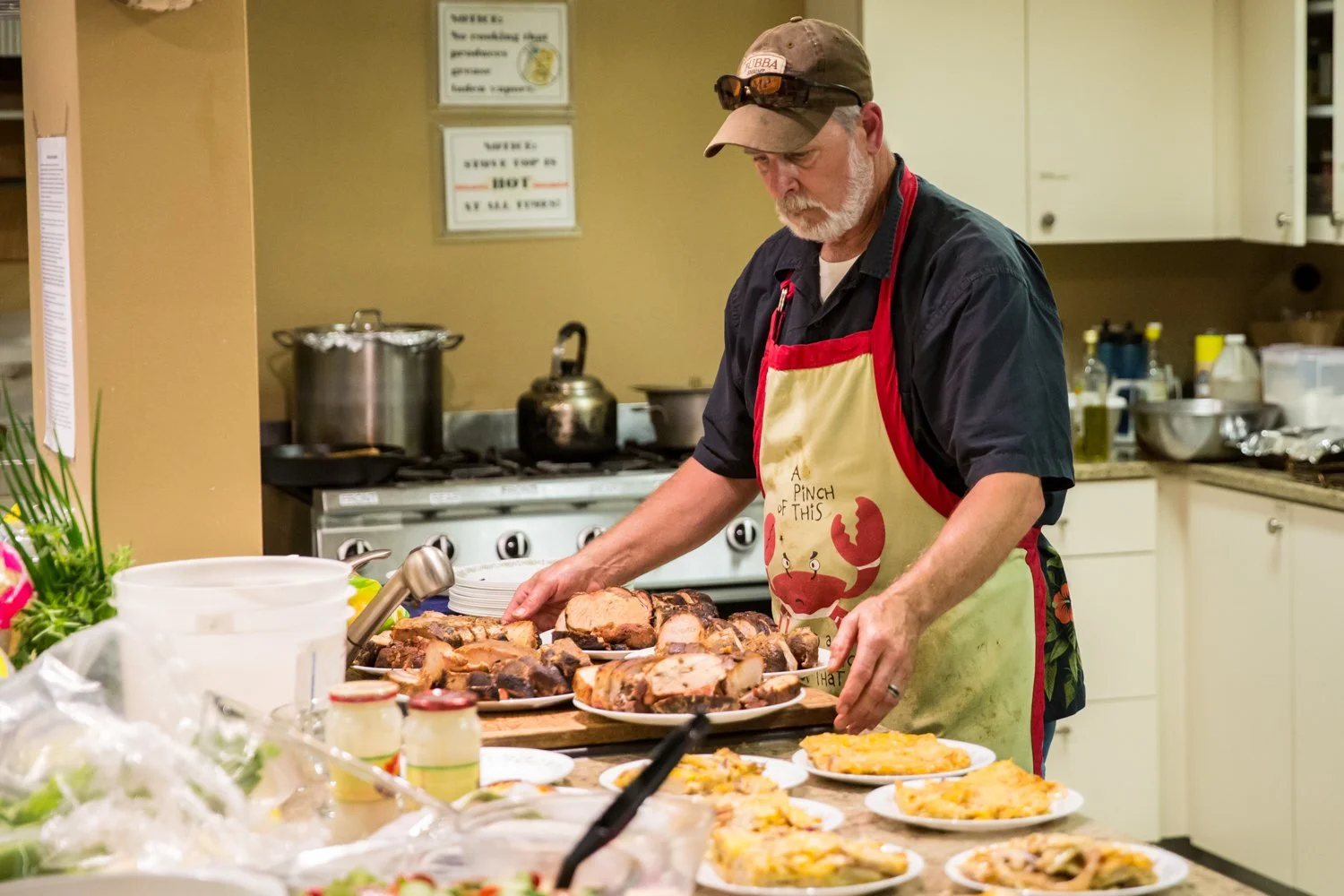 A man wearing a beige cap, black shirt, and an apron with a lobster design is preparing food in a kitchen with plates of cooked meat and dishes on the counter.