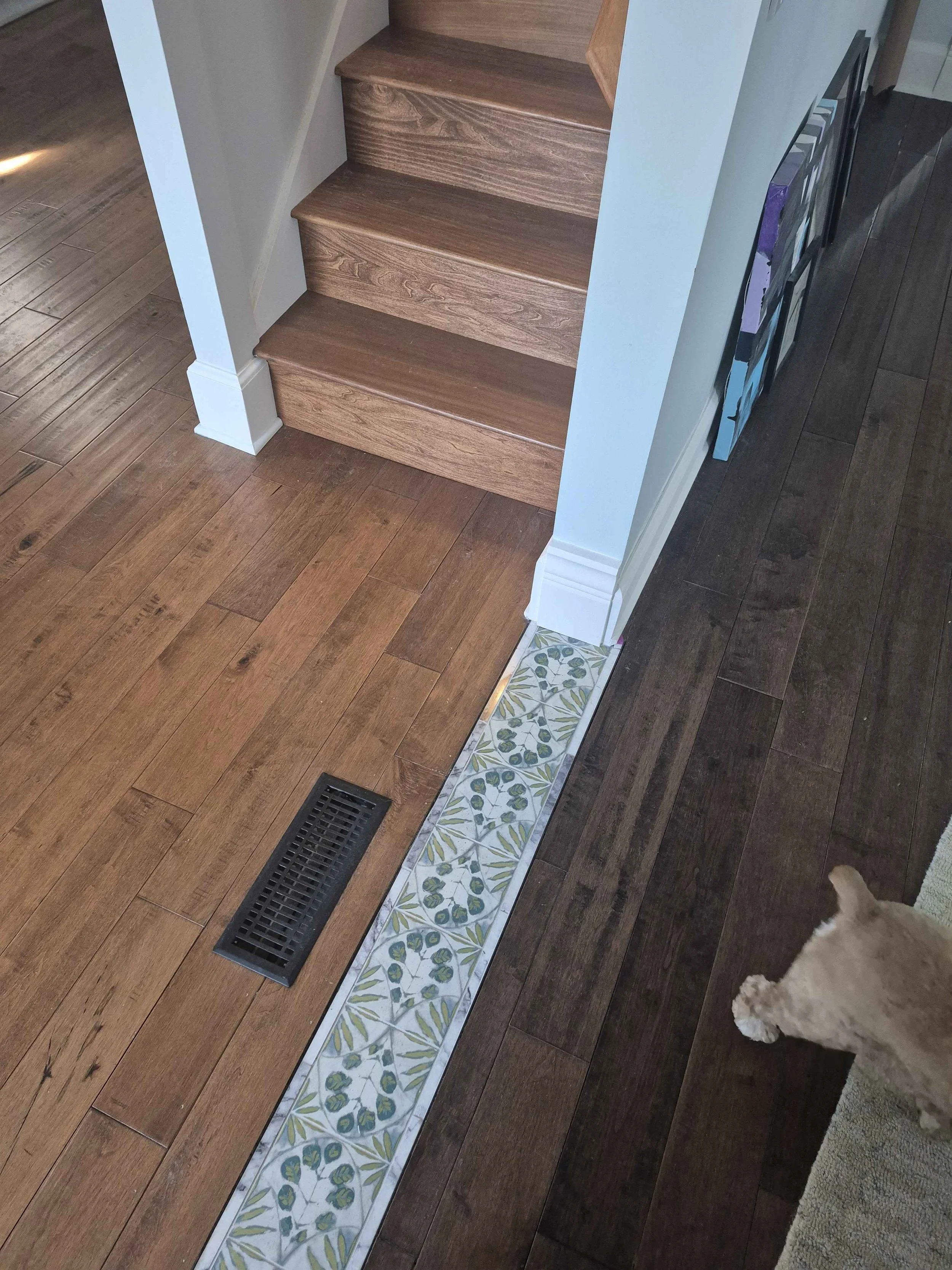 Home flooring transition with decorative tile inlay between two hardwood floors. The image showcases warm-toned wood planks, a patterned tile border with a floral and vine design, and a floor vent. Wooden stairs with white trim lead to an upper level