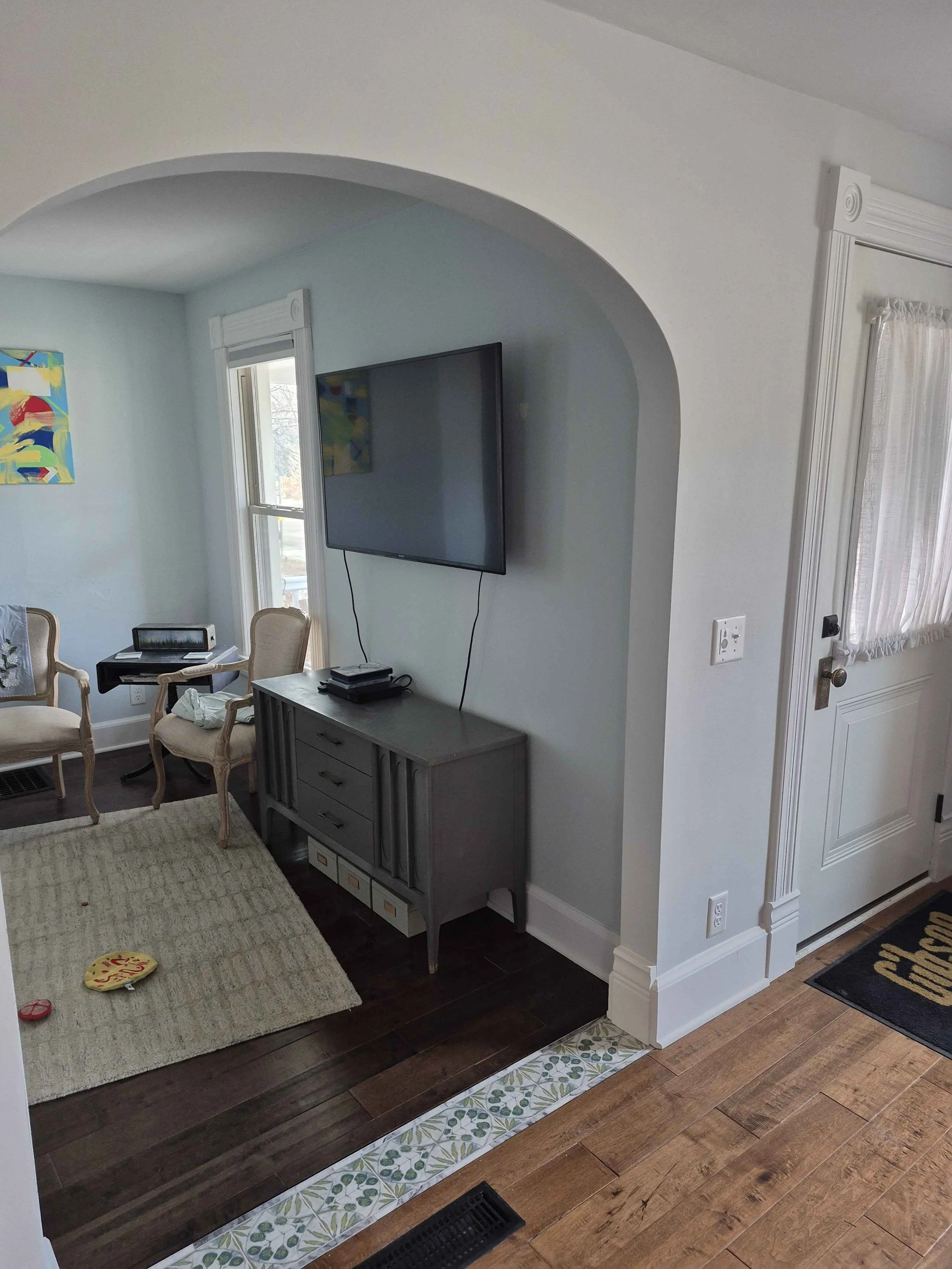 Living room corner with a wall-mounted TV, a small cabinet, and two armchairs on a beige rug. A doorway leads into this space, and there is a piece of modern artwork on the wall. A small decorative item is on the floor.