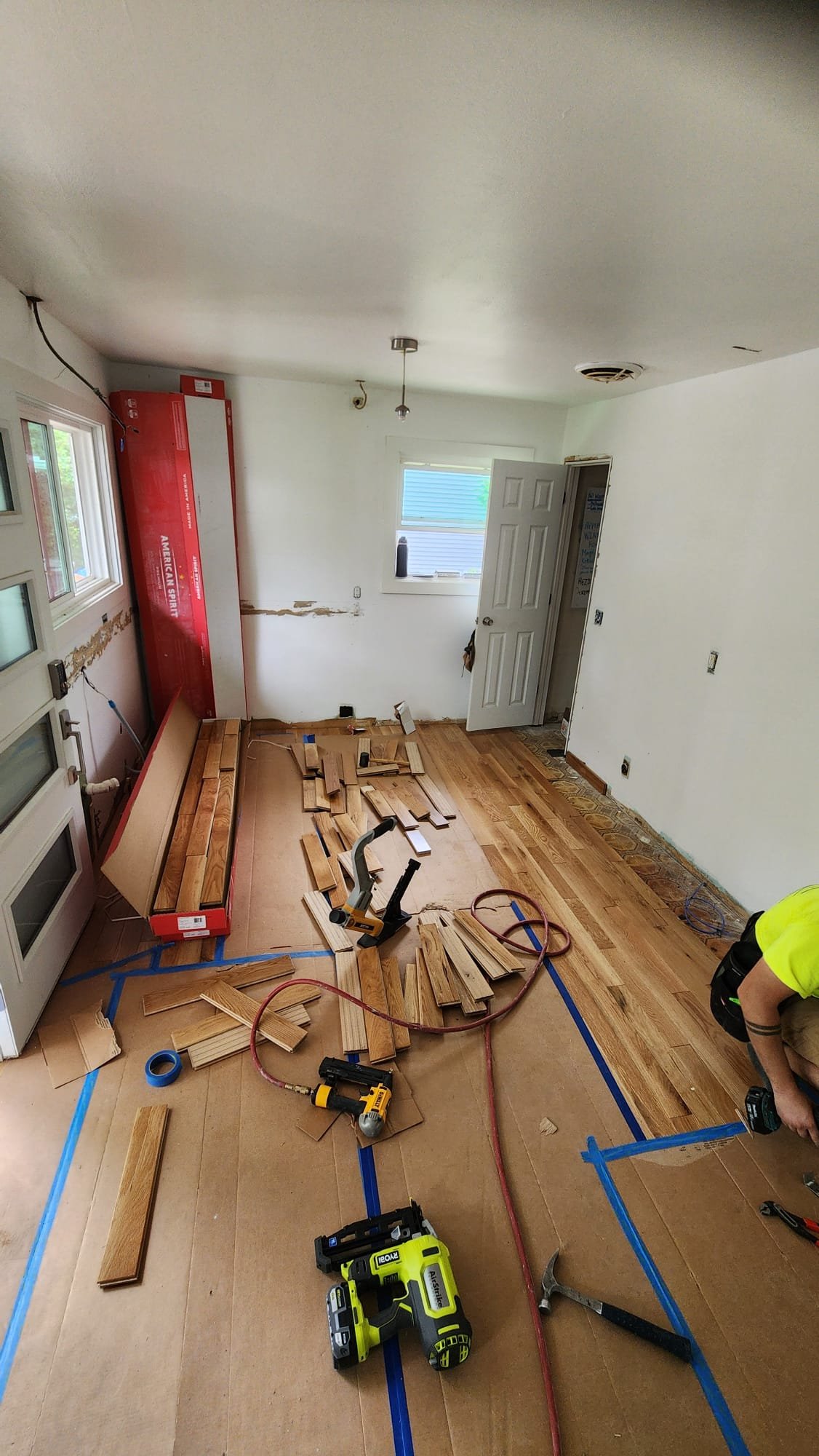 Renovation scene showing a partially installed wooden floor in a room, with tools and wood planks scattered. A person is working in the bottom right.