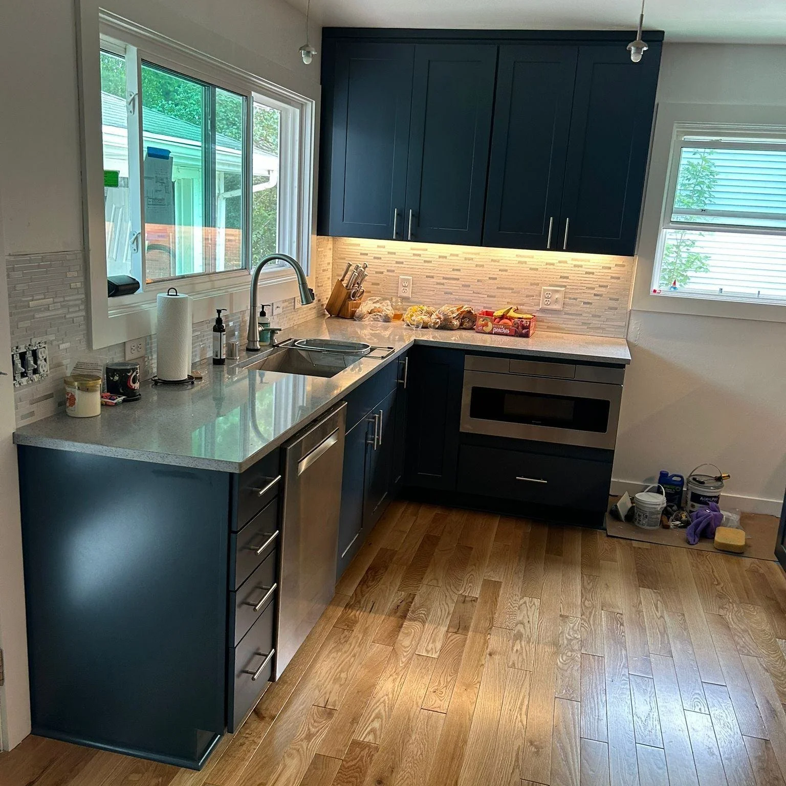 Modern kitchen with dark cabinets, stainless steel appliances, and wooden flooring. The countertop has a sink, paper towels, soap dispenser, and various breads. Wall tiles are light-colored. Natural light comes through a window.