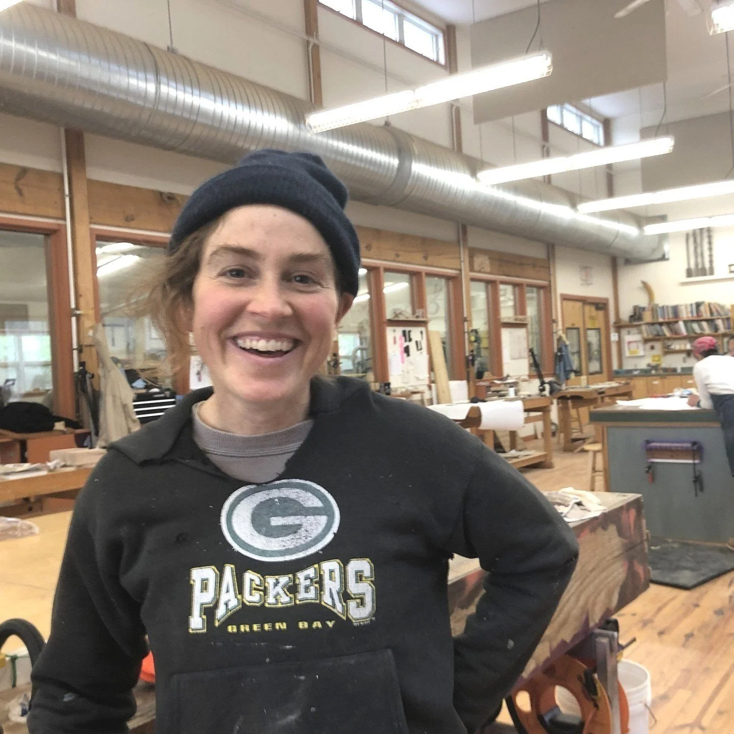The image shows a woodworker wearing a black "Packers Green Bay" sweatshirt, a beanie, standing in a well-lit woodworking shop. They are surrounded by workbenches, woodworking tools, and materials, with other people visible in the background. 