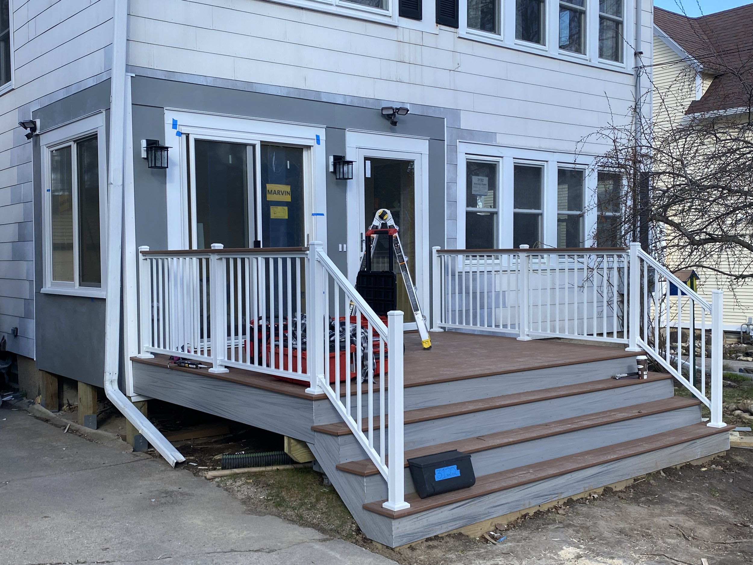 Newly constructed brown and gray porch with white railing on a two-story house, featuring sliding doors, outdoor lights, and tools on the steps.