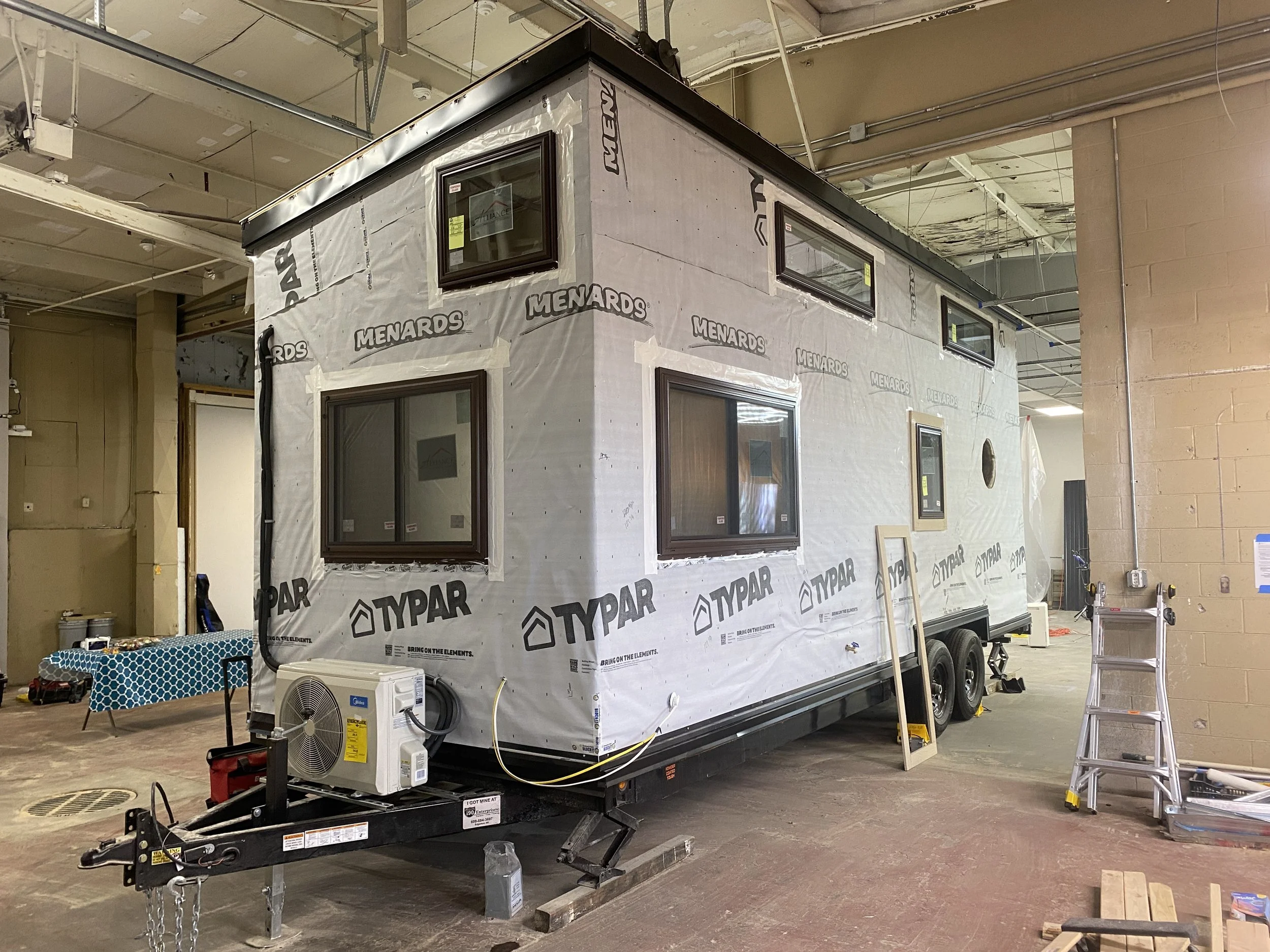 A tiny house under construction in a workshop, wrapped in Typar house wrap with visible windows and an air conditioning unit mounted on the front. The structure is mounted on a trailer for mobility.
