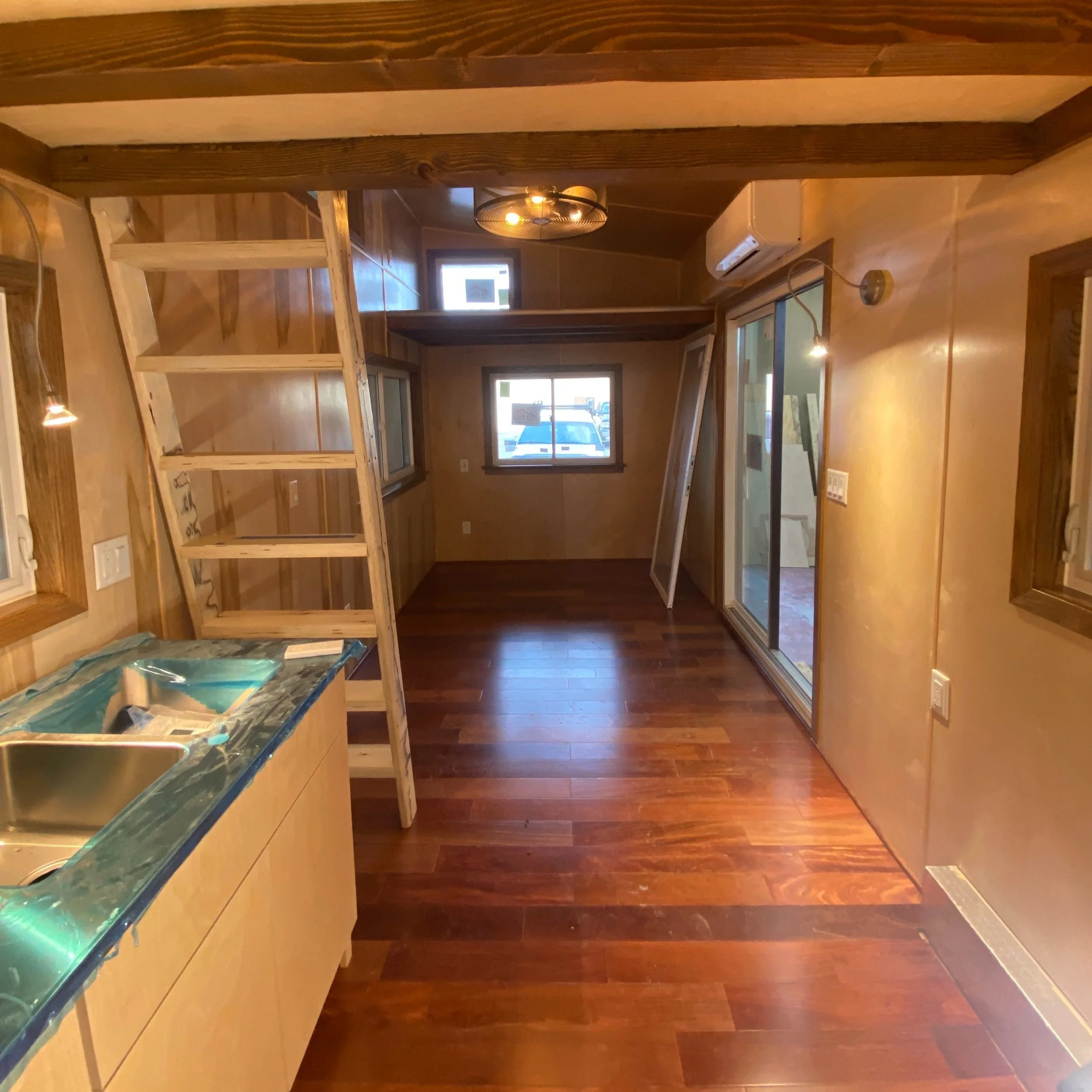Interior of a tiny house featuring a wooden loft ladder, hardwood floors, a small kitchen area with a sink, recessed lighting, multiple windows, and a sliding glass door.