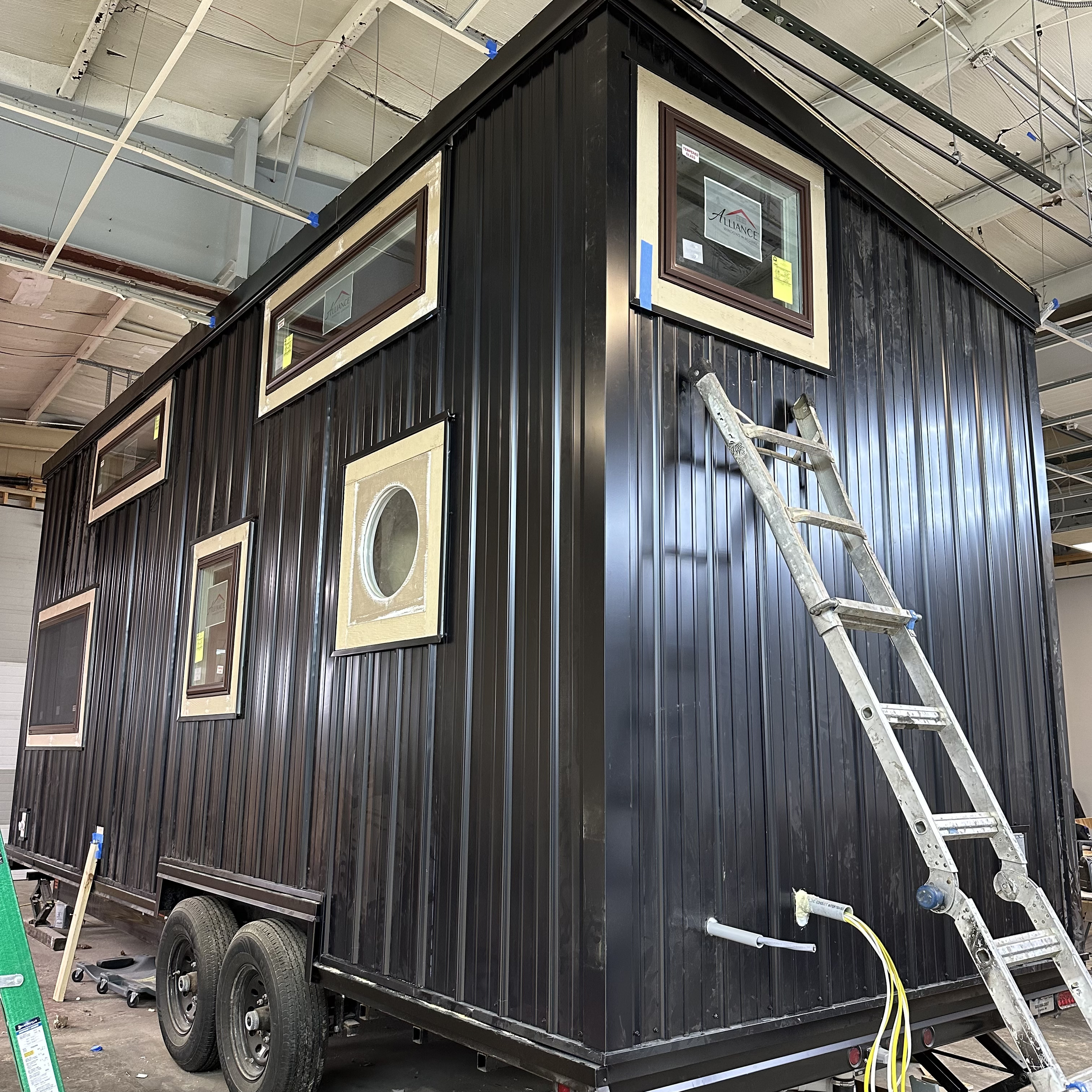 Tiny house under construction with black metal siding, several windows, on a trailer with a ladder leaning against it, inside a workshop.