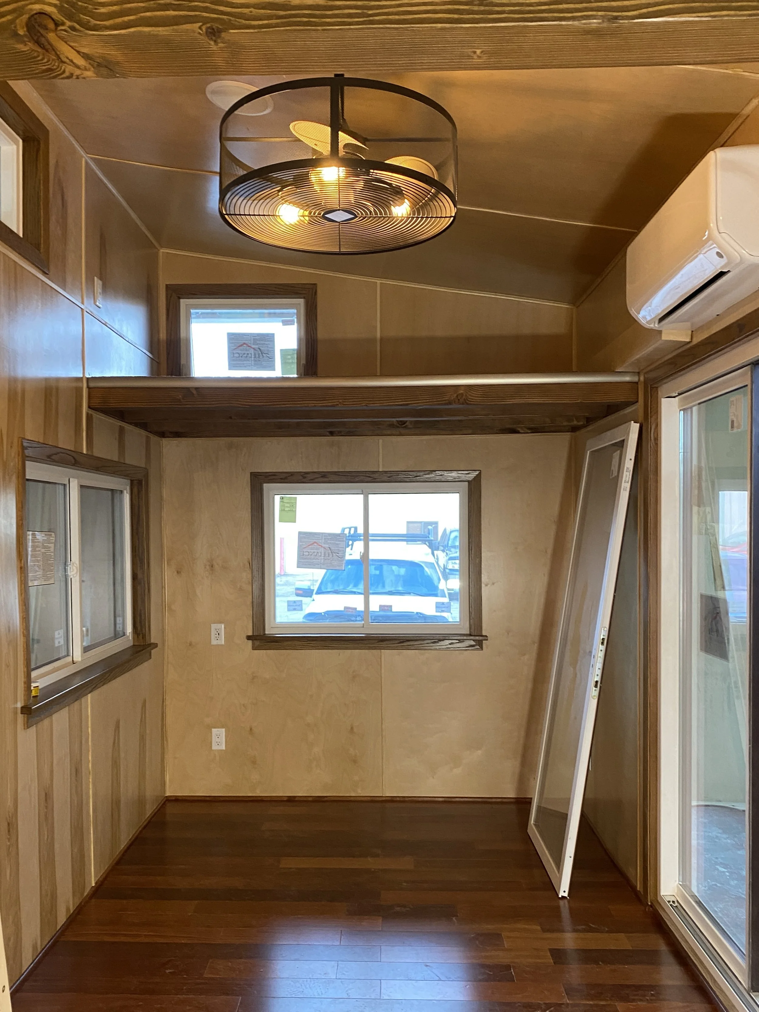 Interior of a wooden tiny house with loft, ceiling light, air conditioner, and multiple windows.