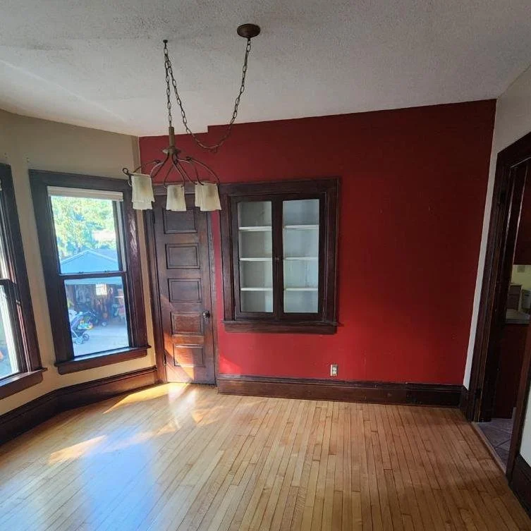 Empty room with hardwood floor and red accent wall, featuring a wooden door, windows, and light fixture.