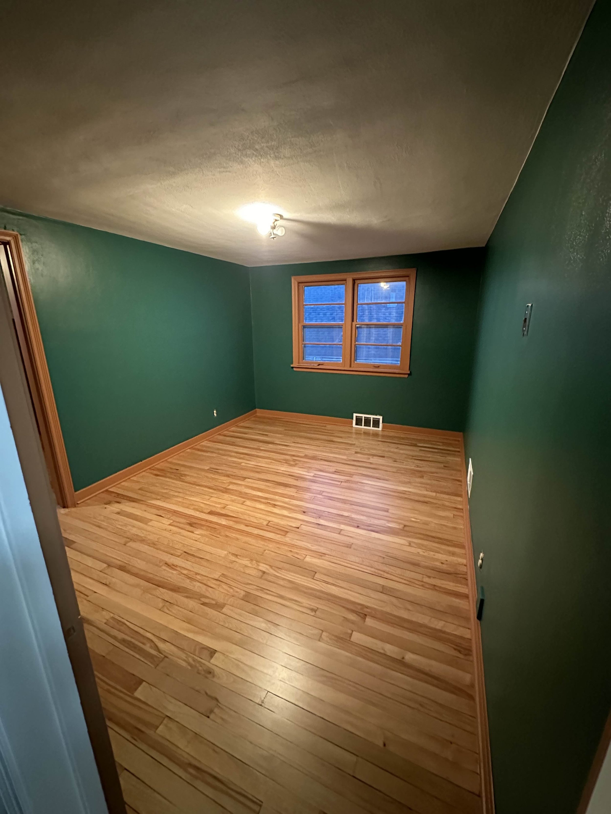 Empty room with wooden floor, green walls, wooden trim, ceiling light, and a window with blue curtains.