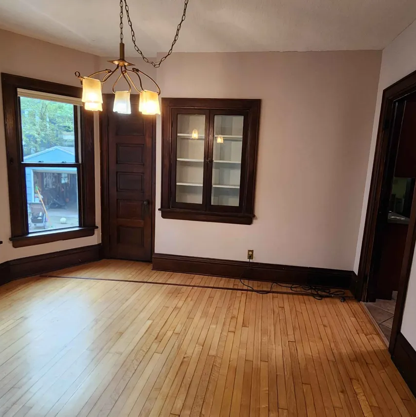 Empty room with hardwood floor, dark wood trim, built-in cabinet, window, door, and hanging light fixture.
