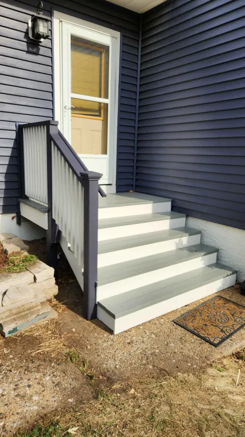 Outdoor porch steps leading to a white door with a glass center. The house has dark blue siding and a small black lantern mounted on the wall. A welcome mat is placed at the foot of the steps.