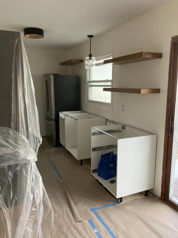 Kitchen under renovation with white cabinets, open shelving, black refrigerator, and plastic coverings on surfaces.