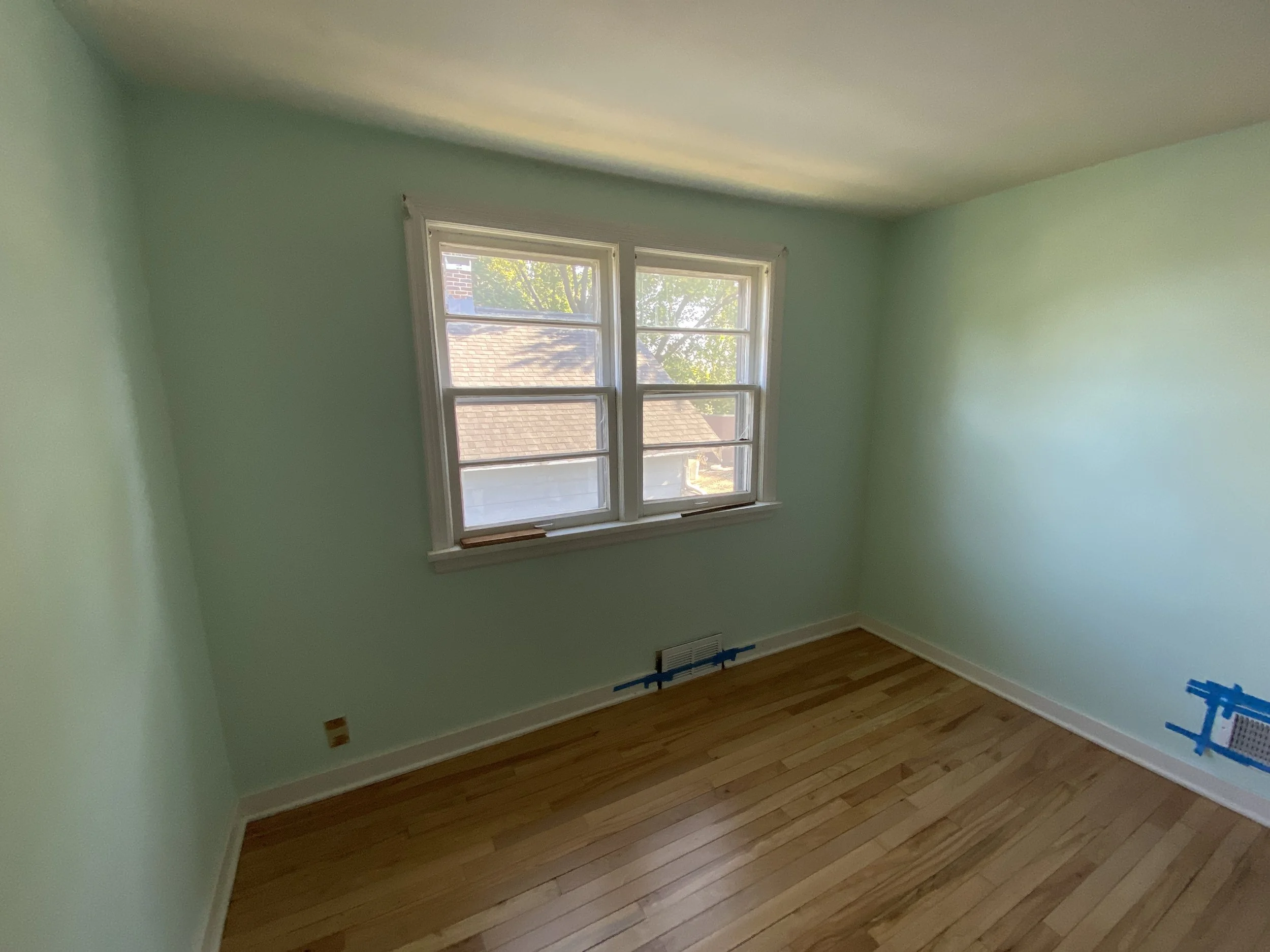 Empty room with light green walls, a wooden floor, and a window facing outdoors.