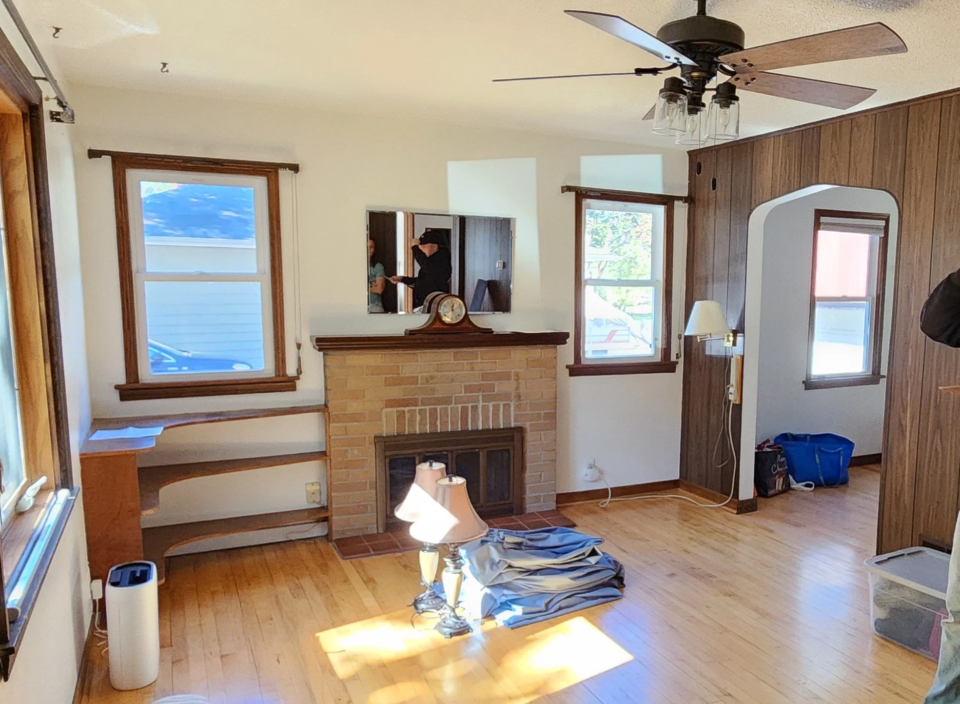 Living room with a ceiling fan, wooden flooring, brick fireplace, two table lamps, a wall mirror, and several windows. There are also some miscellaneous items like a blue bag and a box.