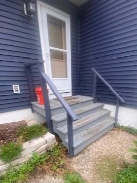 Outdoor porch with steps and railing leading to a white door, surrounded by blue siding and a small garden area.