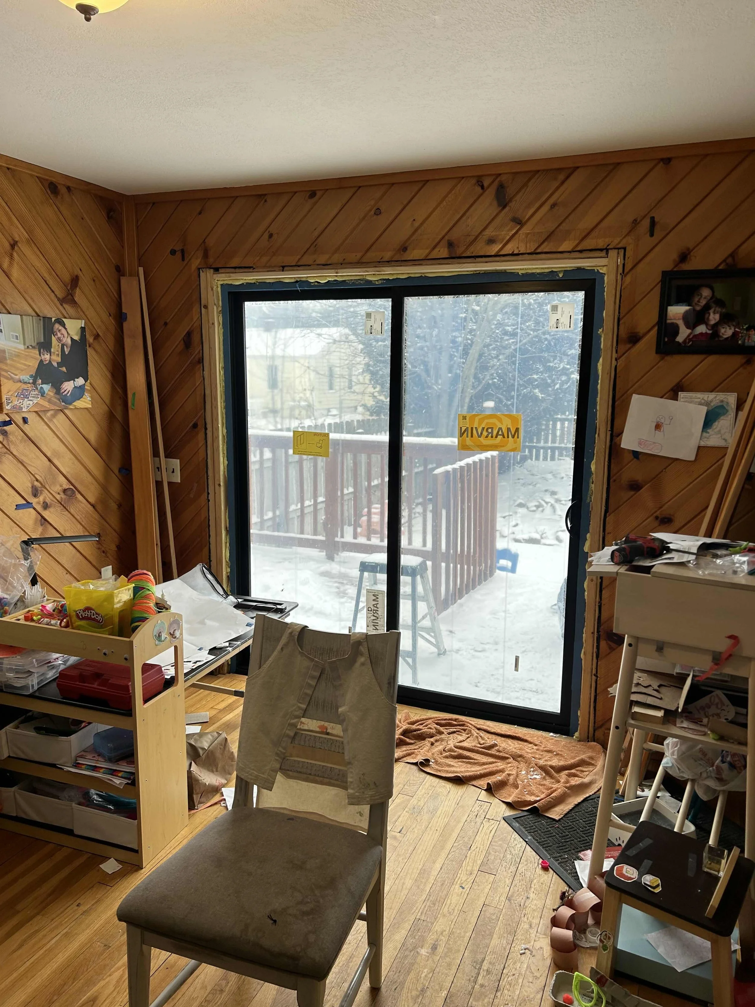 Interior room with wooden walls, sliding glass door leading to snowy deck, children's artwork, and cluttered furniture and toys.