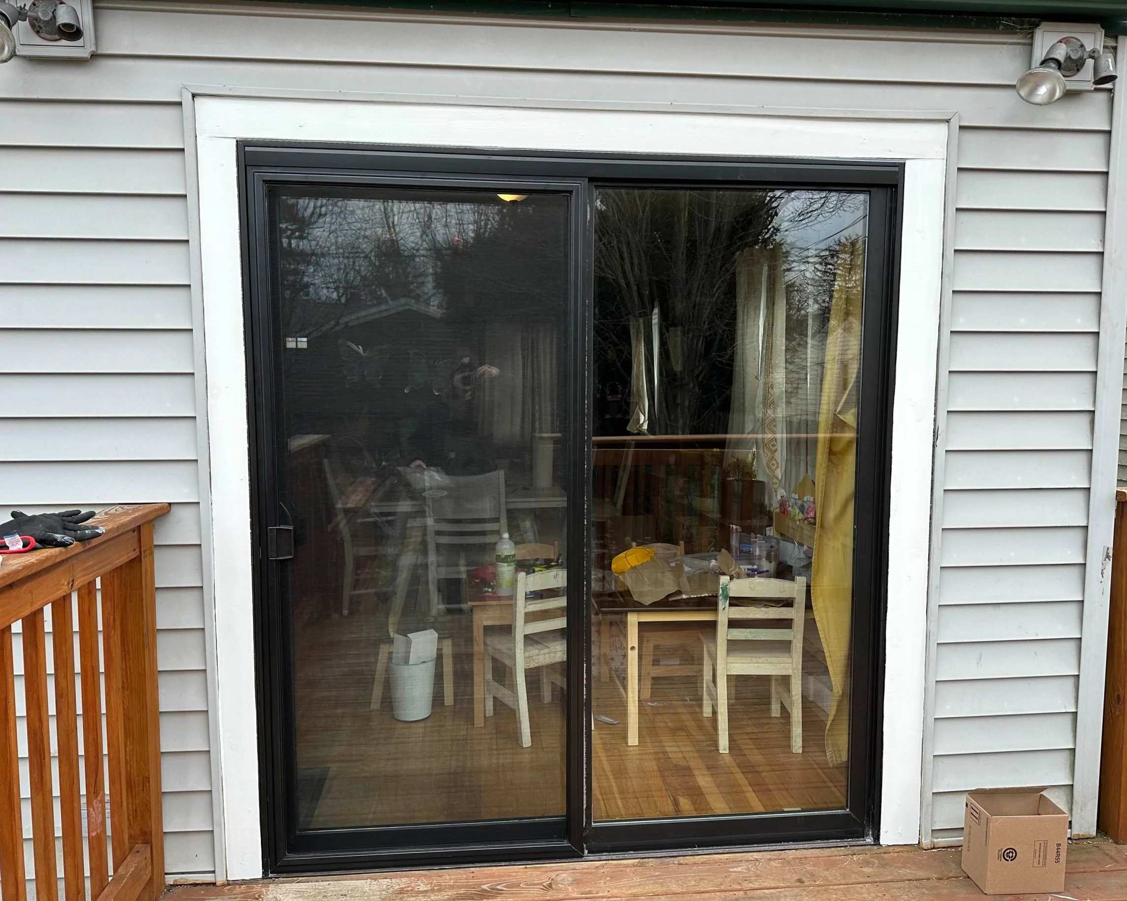 Black-framed sliding glass patio door with white trim on gray vinyl siding, leading to a wooden deck and interior kitchen with children's table set