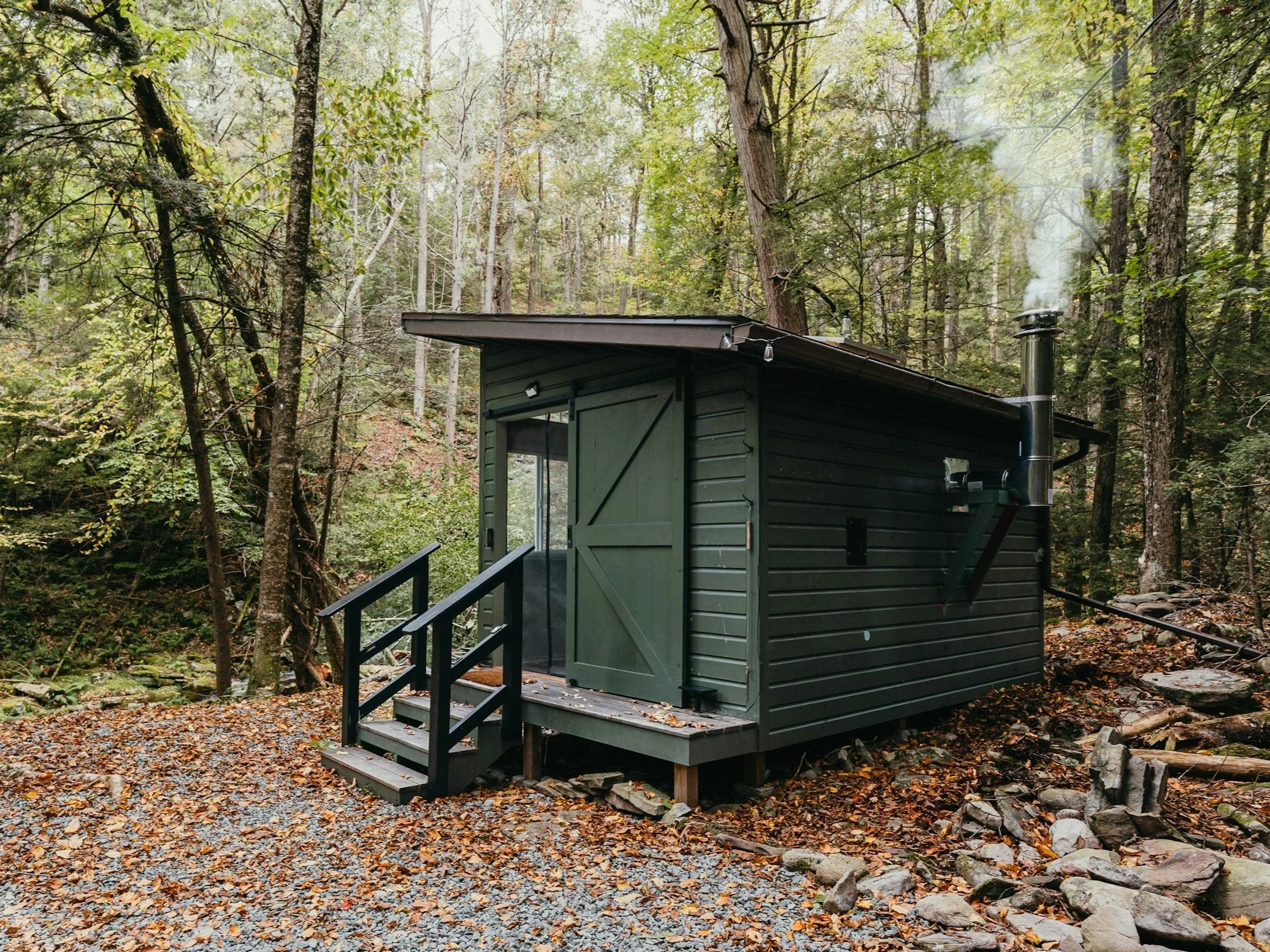 Charming, secluded cabin-style sauna in a peaceful forest retreat, featuring a cozy chimney and surrounded by scenic natural beauty with fall leaves and tall trees.