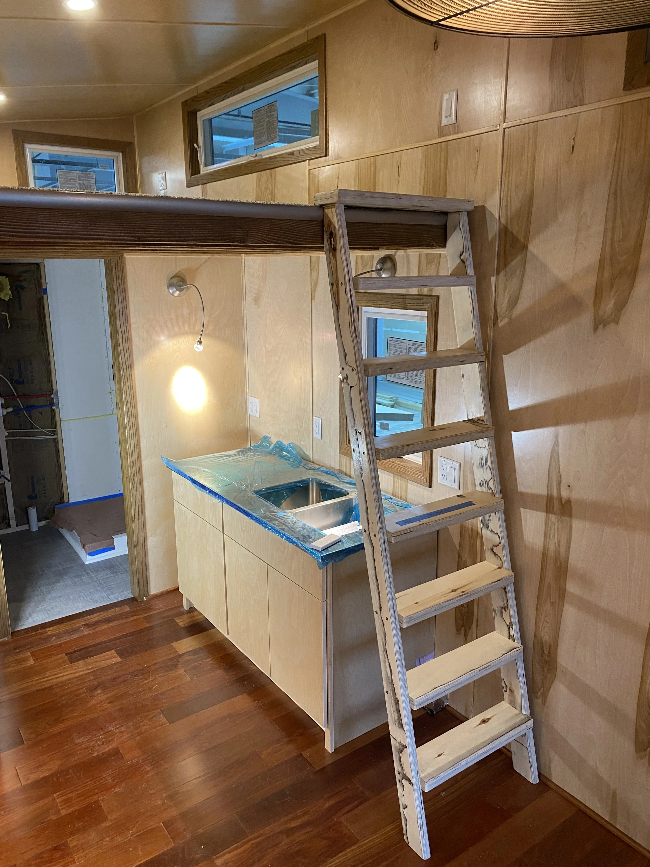 Interior of a small modern wooden room with a wooden ladder leading to a loft space. There is a kitchenette with a sink, countertop, and light fixtures. The floor is made of hardwood.