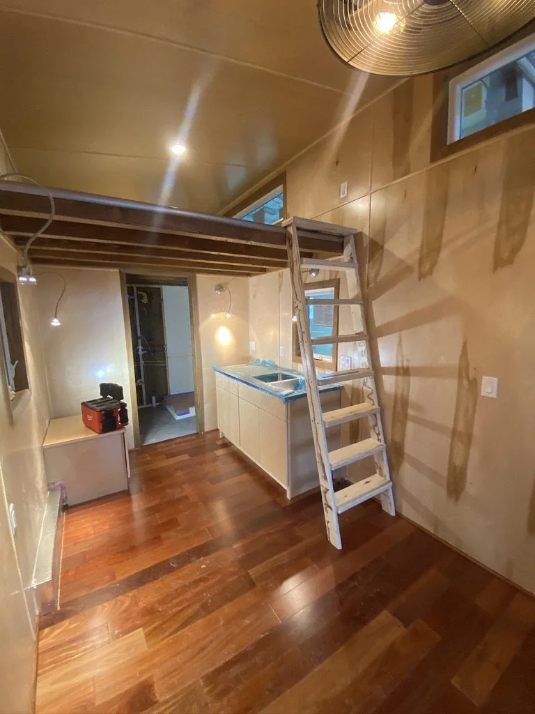 Interior of a tiny house with wooden floors, a ladder leading to a loft, a small kitchenette area, and exposed lighting.