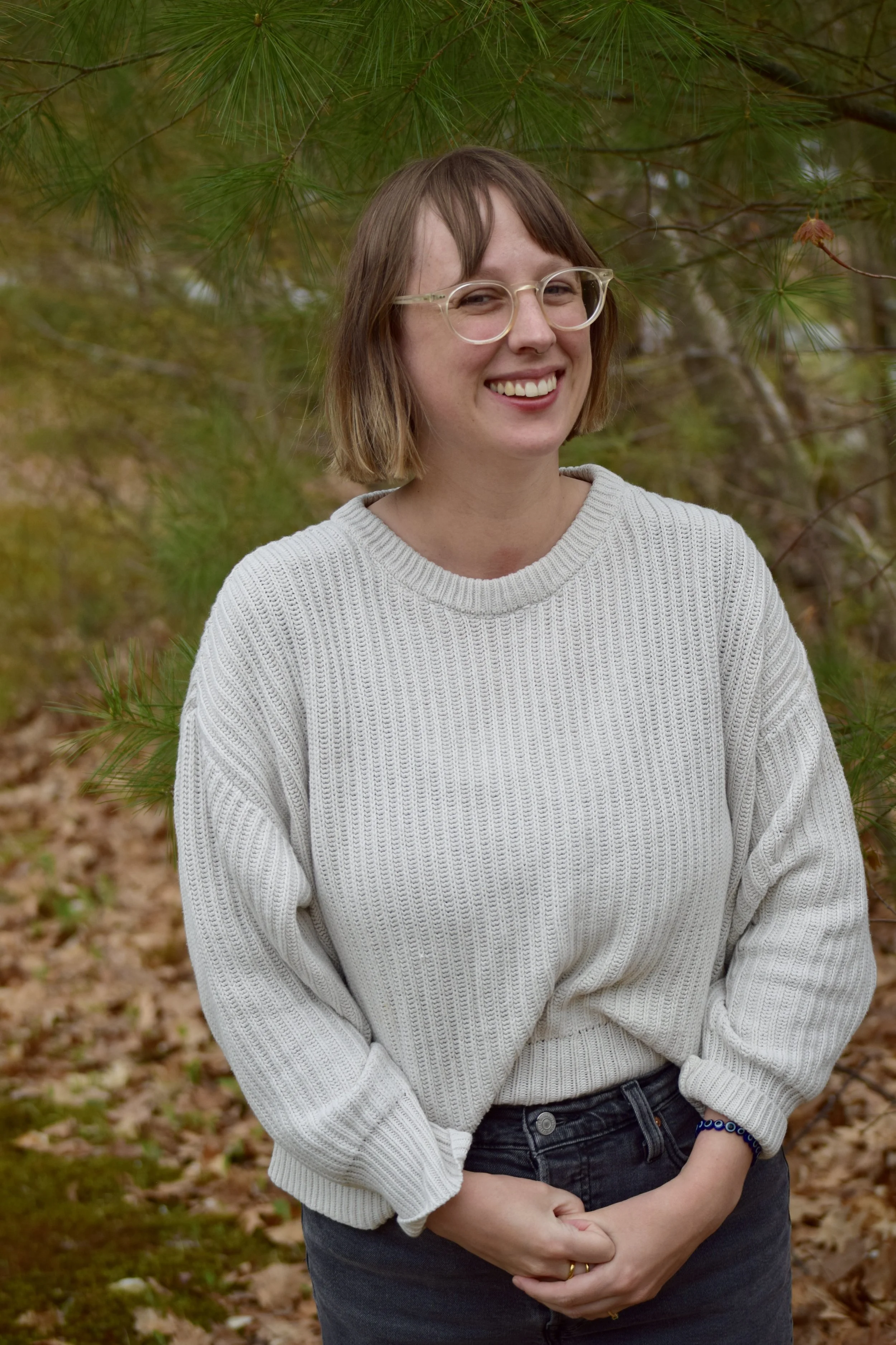 A woman with short brown hair, wearing glasses, a light gray sweater, and dark jeans, standing outdoors among trees with fallen leaves on the ground, smiling at the camera.