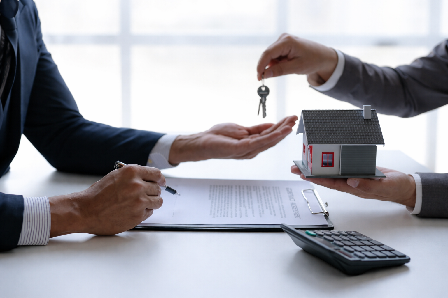Two people in business suits exchanging keys over a small house model during a home transaction or real estate deal.