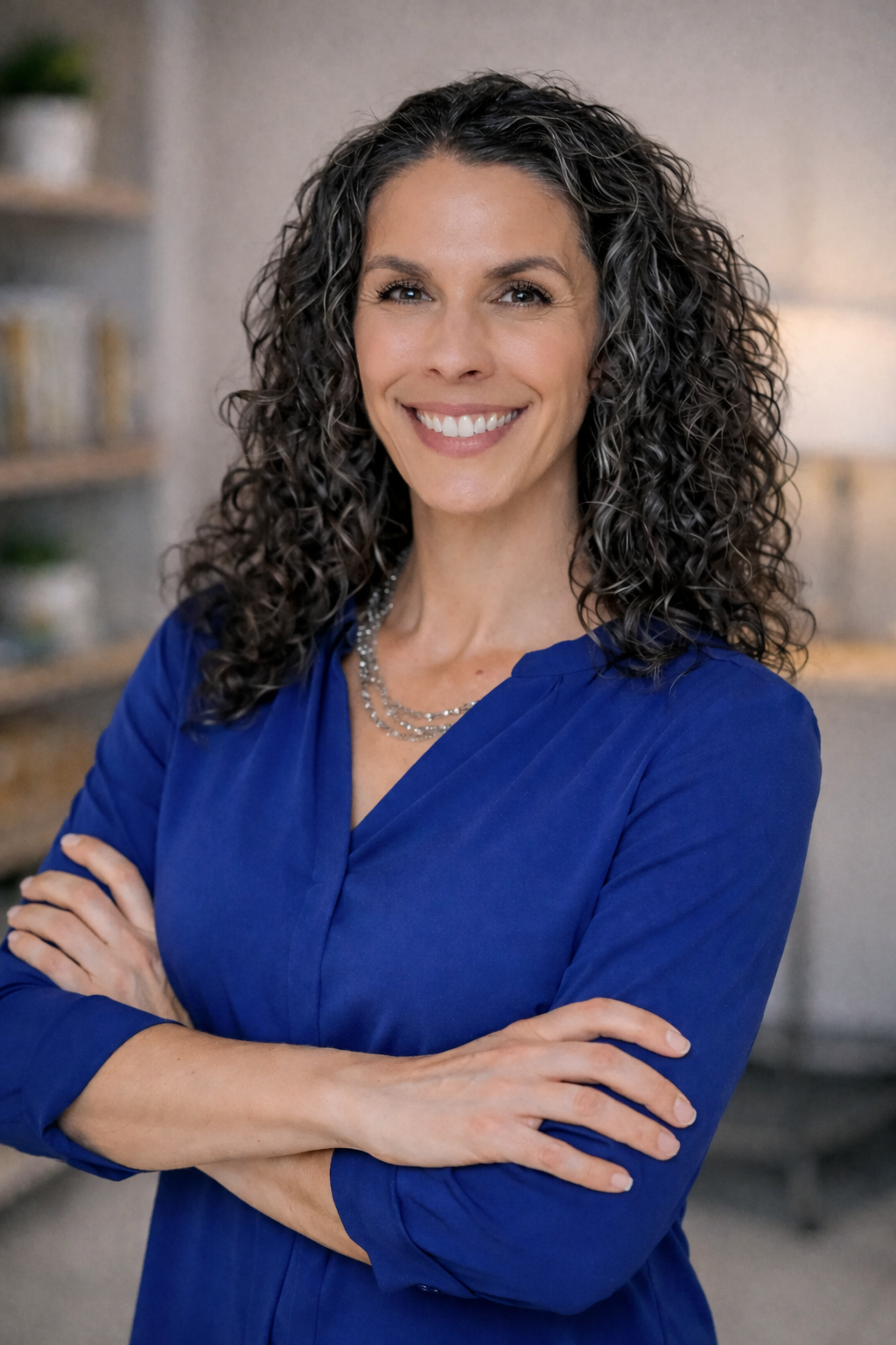 Danielle Jefferies, A woman with curly dark hair smiling, wearing a blue blouse and layered necklaces, standing with arms crossed in an indoor setting.