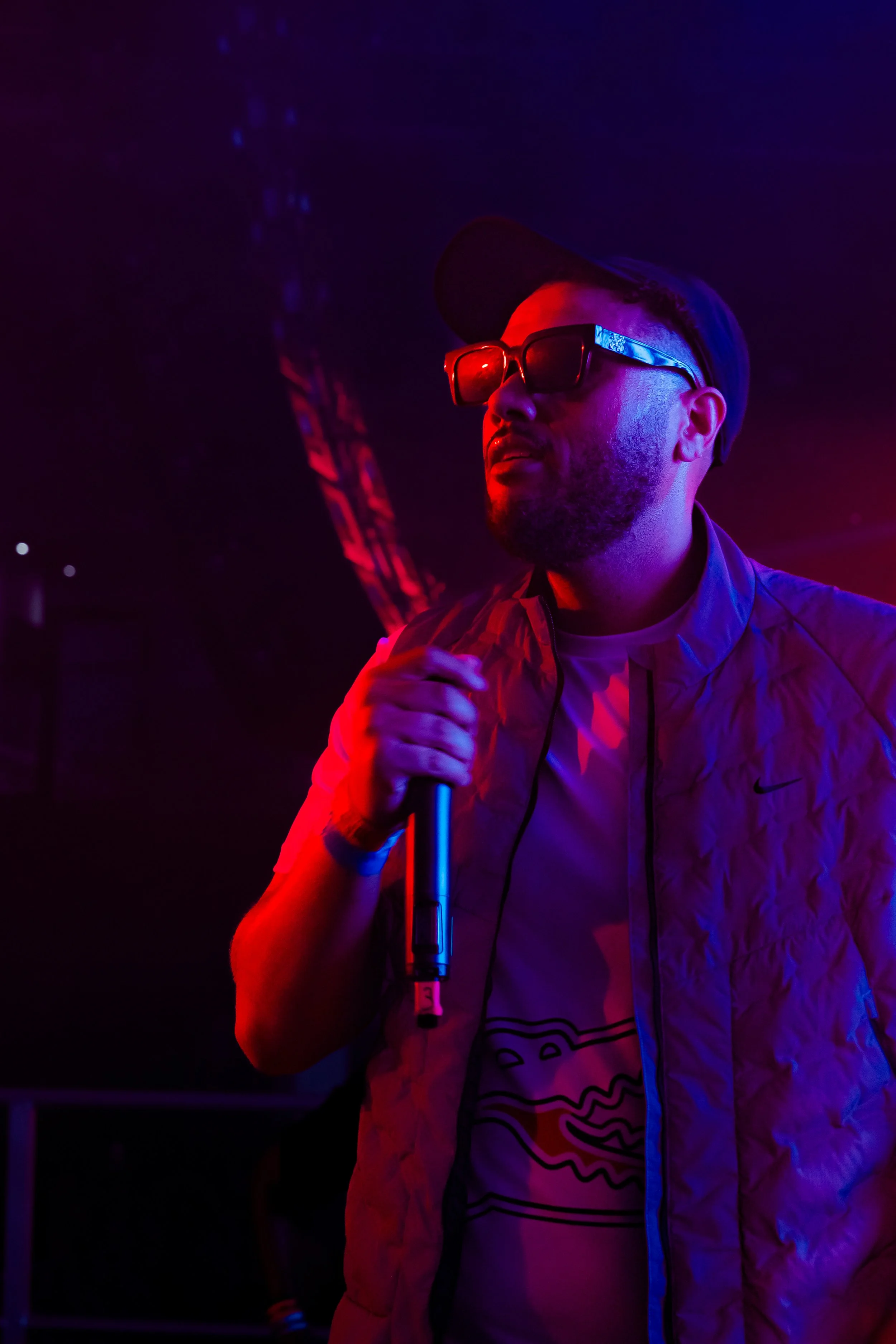 A man with a beard wearing sunglasses and a backwards cap, holding a microphone in a dark, neon-lit setting.