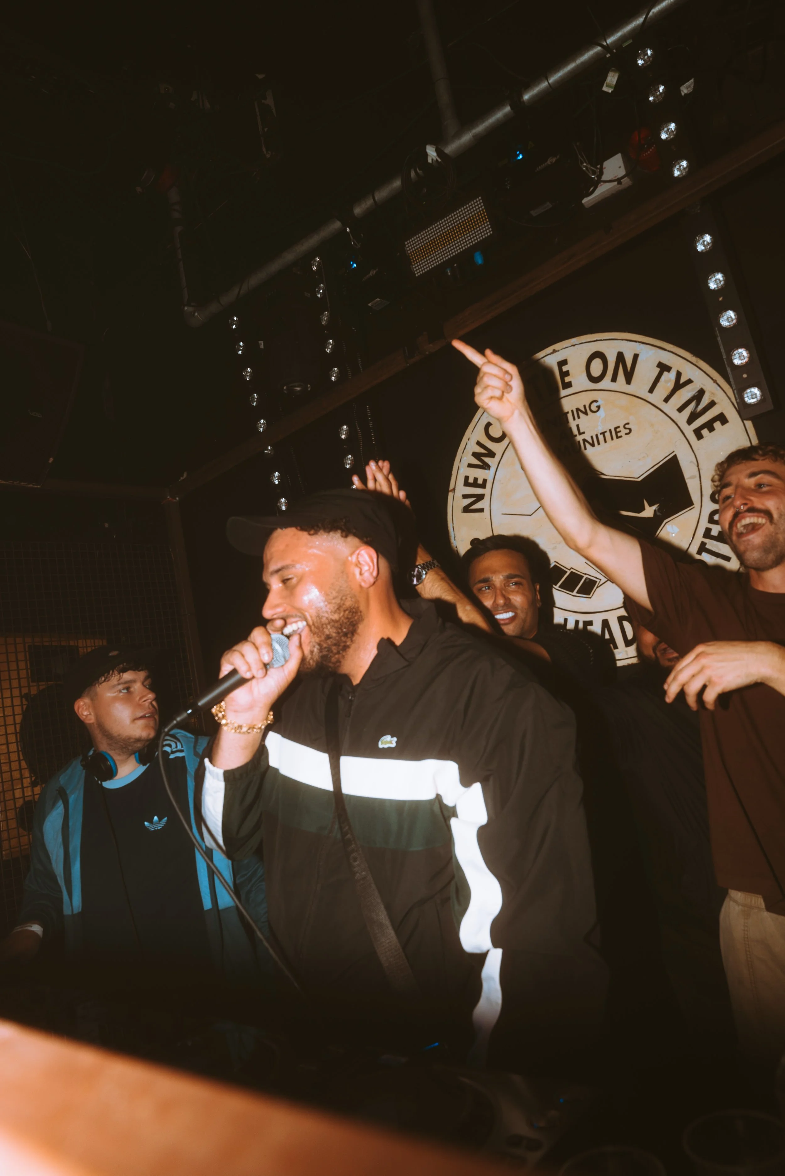 A group of friends celebrating and smiling at a party or nightclub, with one person holding a microphone and wearing a black and white jacket, others raising their hands, and a DJ behind them, against a backdrop with a large circular sign.