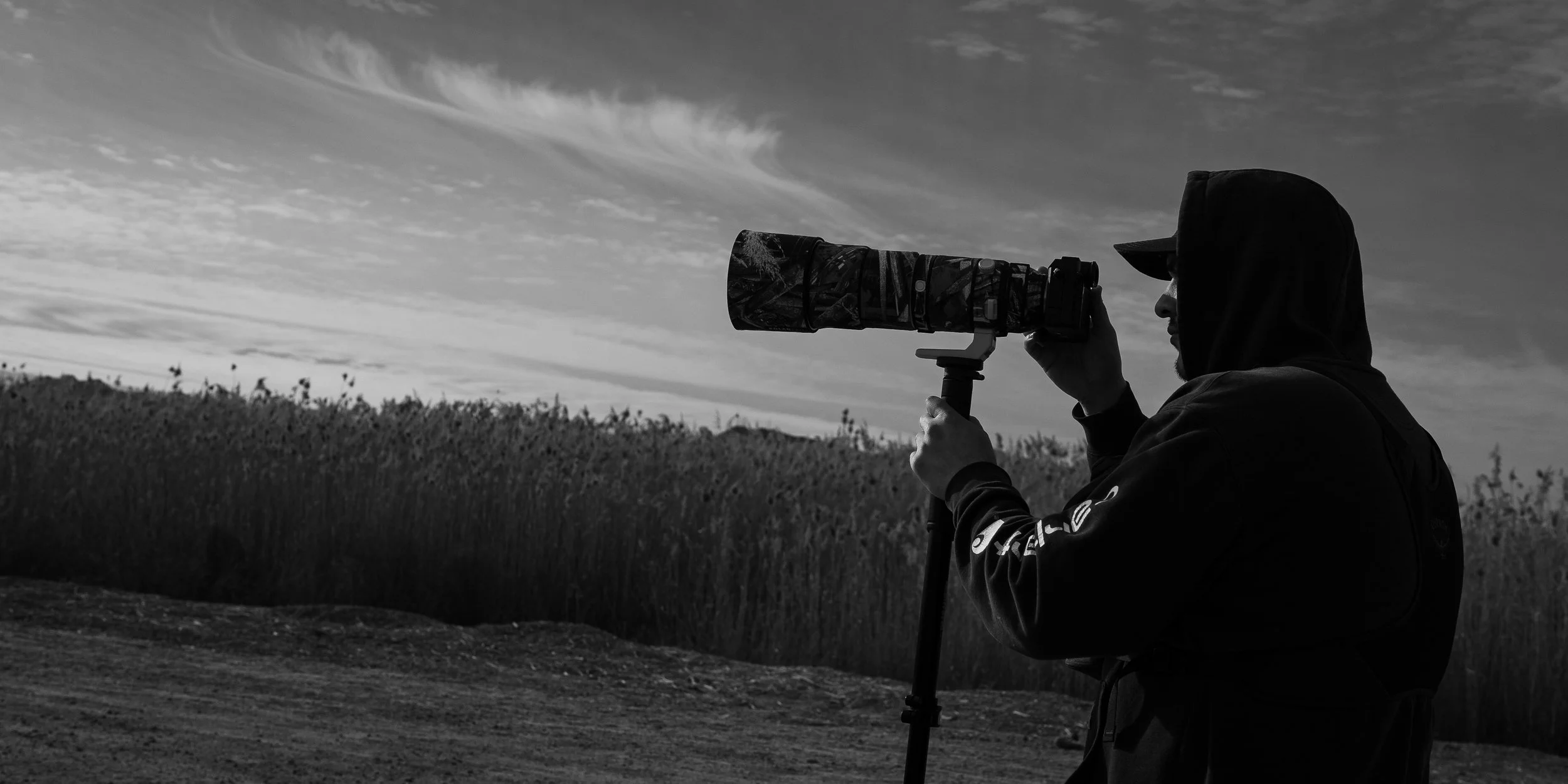 Person dressed in a hoodie using a camera with a telephoto lens, outdoors during daytime, with a cloudy sky and tall grass in the background.