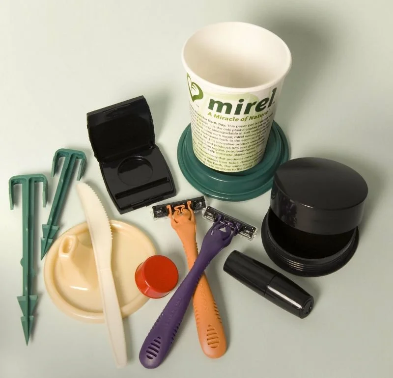 A collection of dental care items including a toothbrush, razor blades, a small black container, a white and red container, a black round container, an empty paper cup on a green saucer, and green dental floss picks.